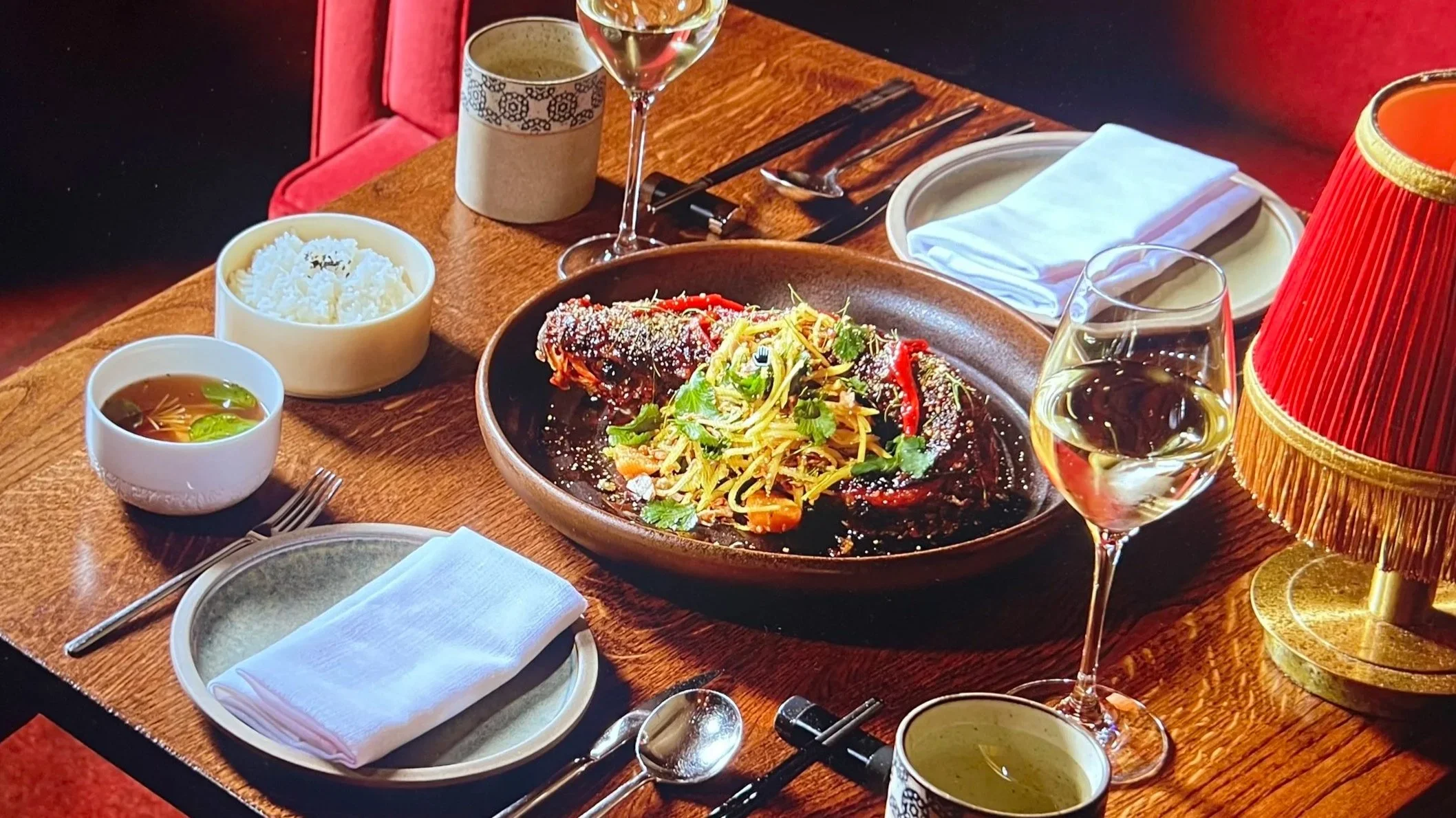 A dining table set with a dish of grilled meat topped with salad, two glasses of white wine, a bowl of rice, a bowl of soup, utensils, a folded napkin, a lamp, and a plate.
