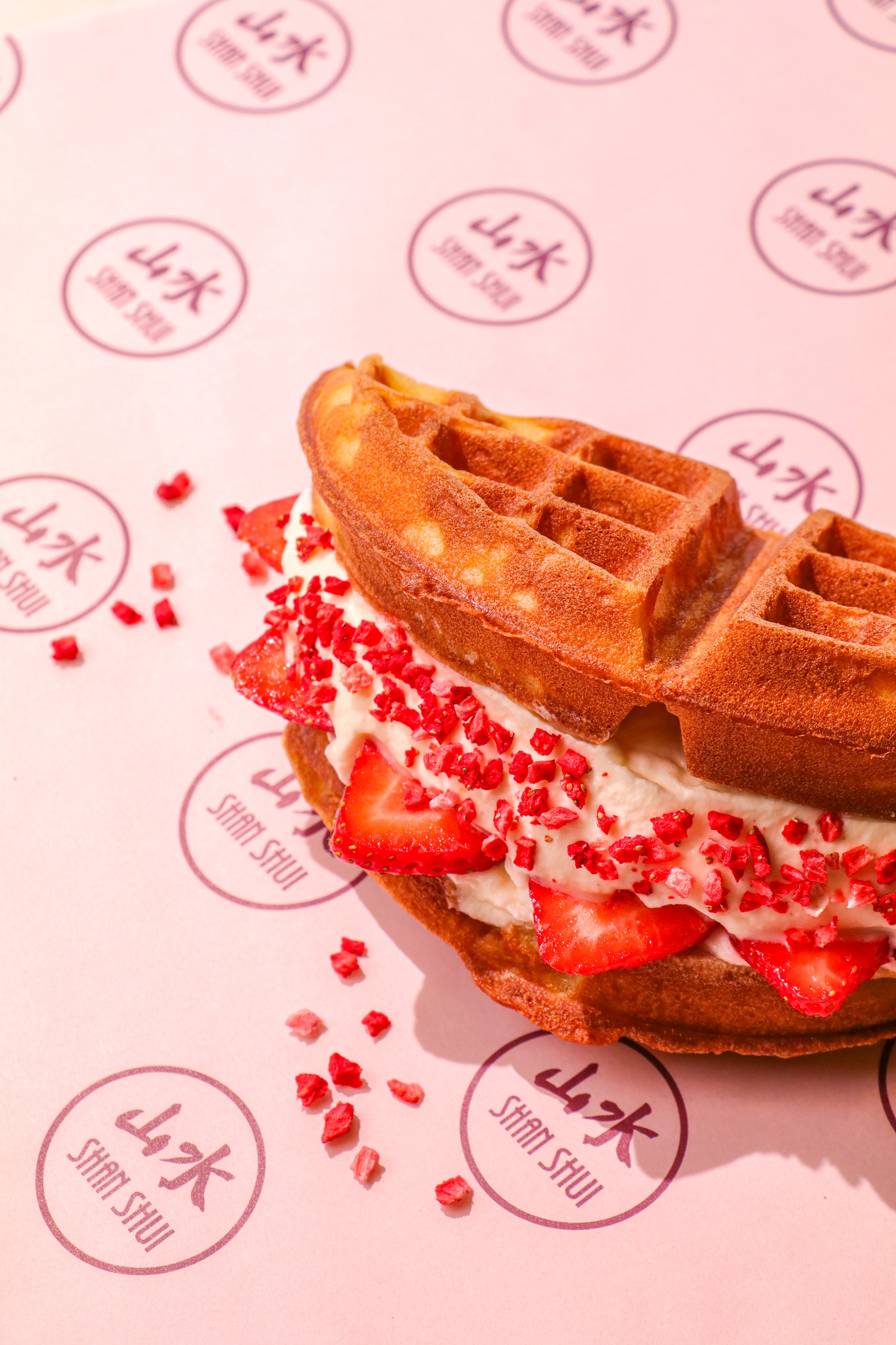 A waffle sandwich filled with strawberries and whipped cream, topped with red sprinkles, placed on pink paper with logos.