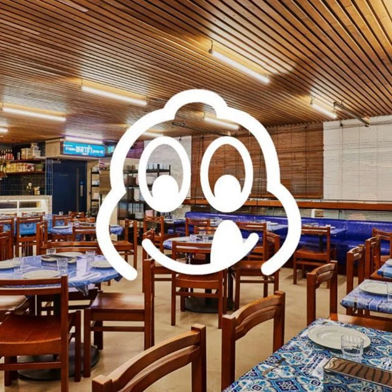 A restaurant interior with wooden chairs, tables with blue tablecloths, and a Michelin Bib Gourmand logo overlaying the image.