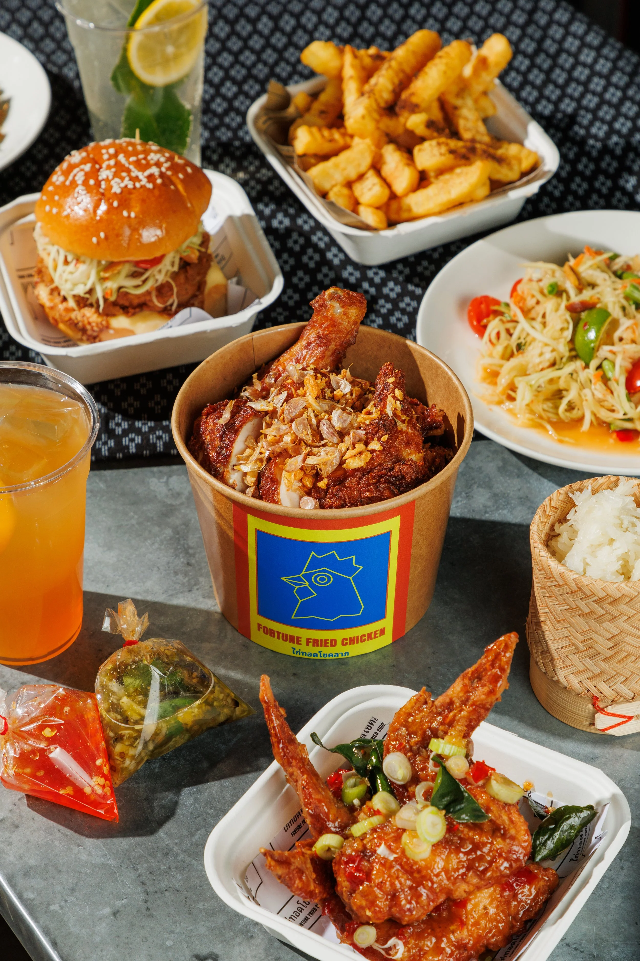 A variety of Asian and American dishes including fried chicken, a burger with coleslaw, French fries, a noodle salad, and drinks on a table.