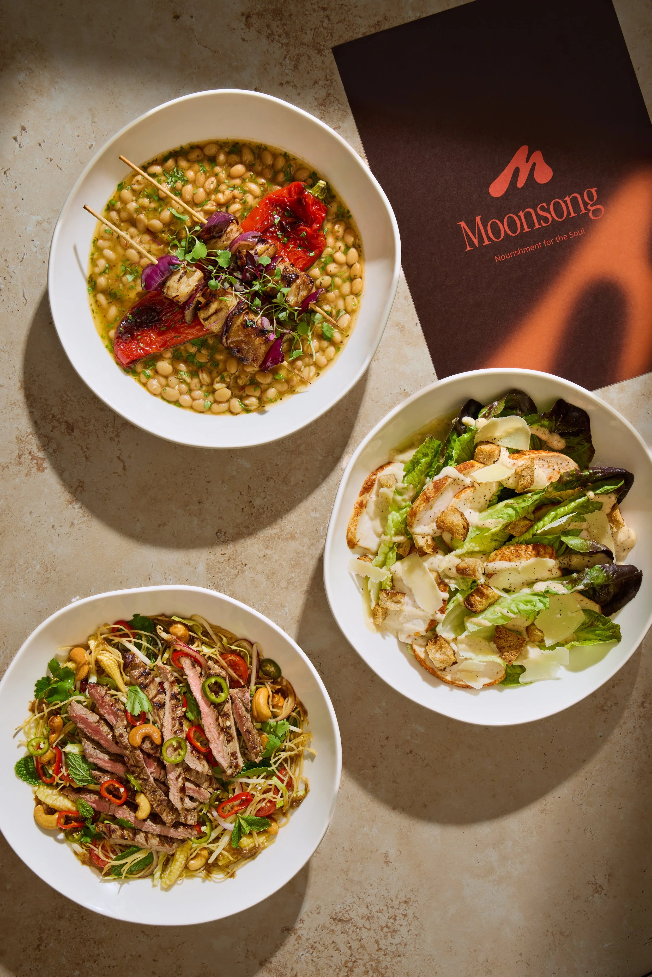 Three bowls of food on a beige surface, with a Moonsong menu nearby. The top bowl contains a bean dish with roasted peppers, eggplant, and microgreens. The right bowl contains a salad with lettuce, chicken, croutons, and shaved cheese. The bottom bowl has a noodle salad with sliced beef, chili, cilantro, and vegetables.