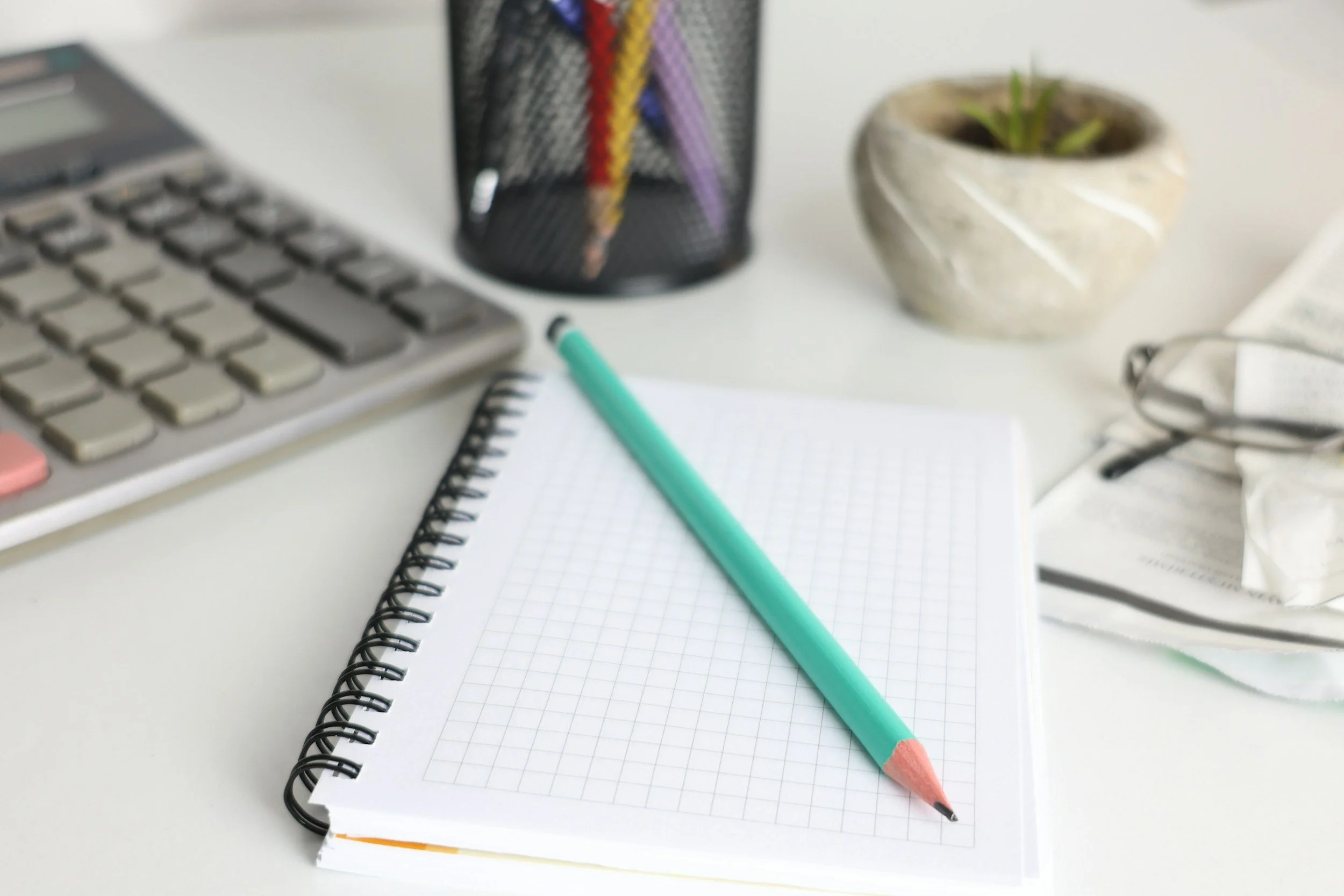Office desk with a notebook, a pen, a calculator, a black mesh pencil holder with colored pencils, a plant in a stone pot, reading glasses, and a crumpled tissue.