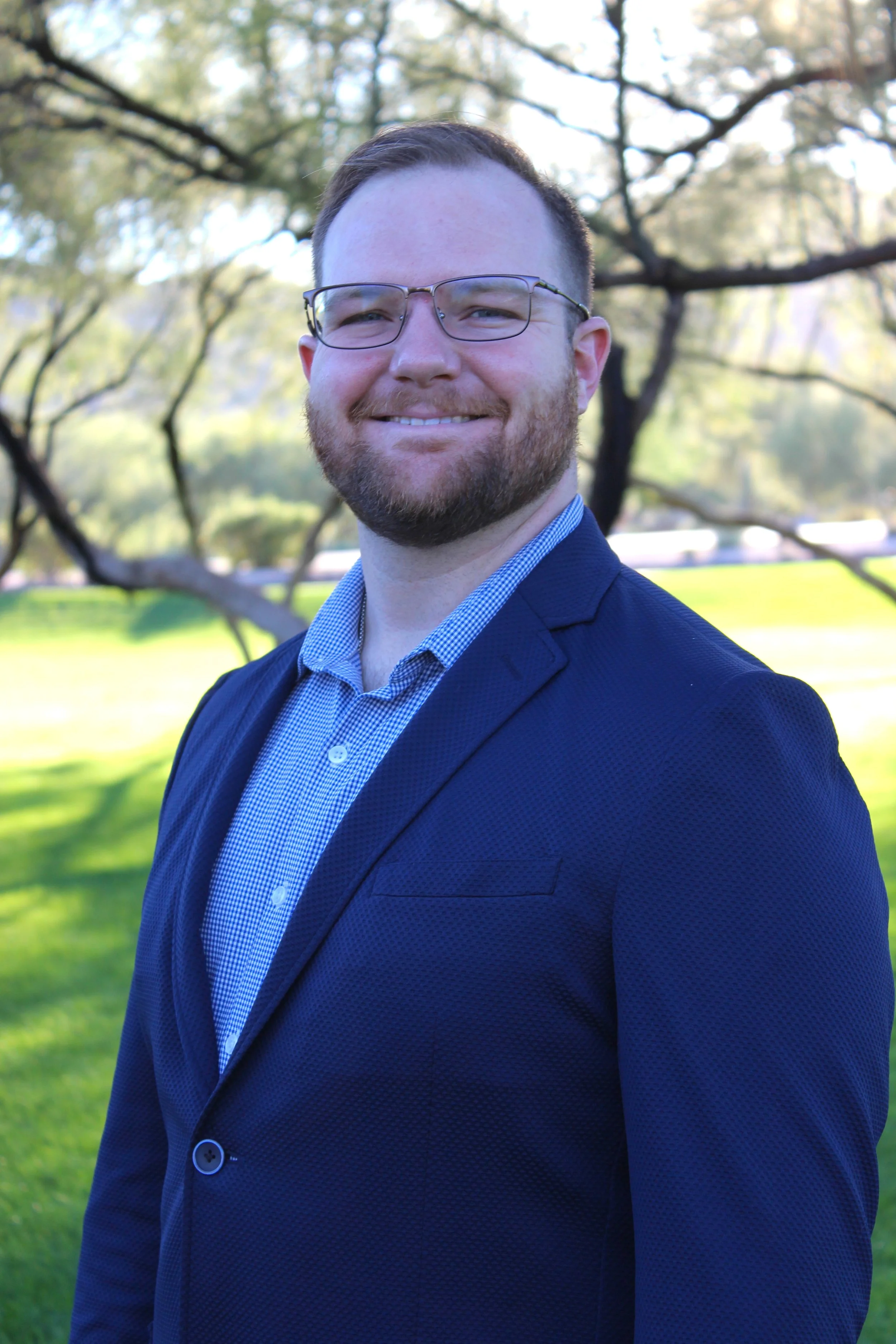 A man with glasses and a beard smiling outdoors in a park, wearing a navy blazer and a patterned button-up shirt, with trees and grass in the background.