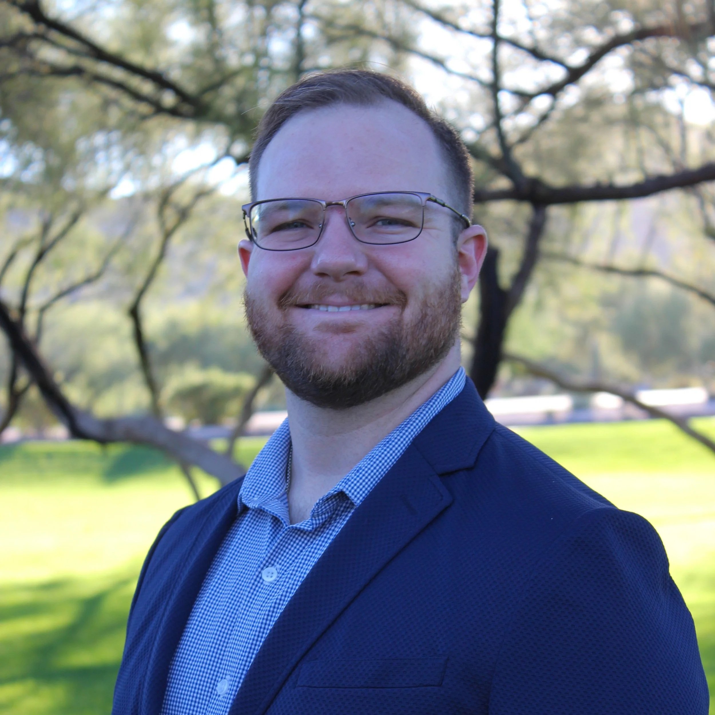 Headshot of a man with glasses and a beard, smiling outdoors during daytime, wearing a dark blazer and blue checkered shirt, with trees and sunlight in the background.