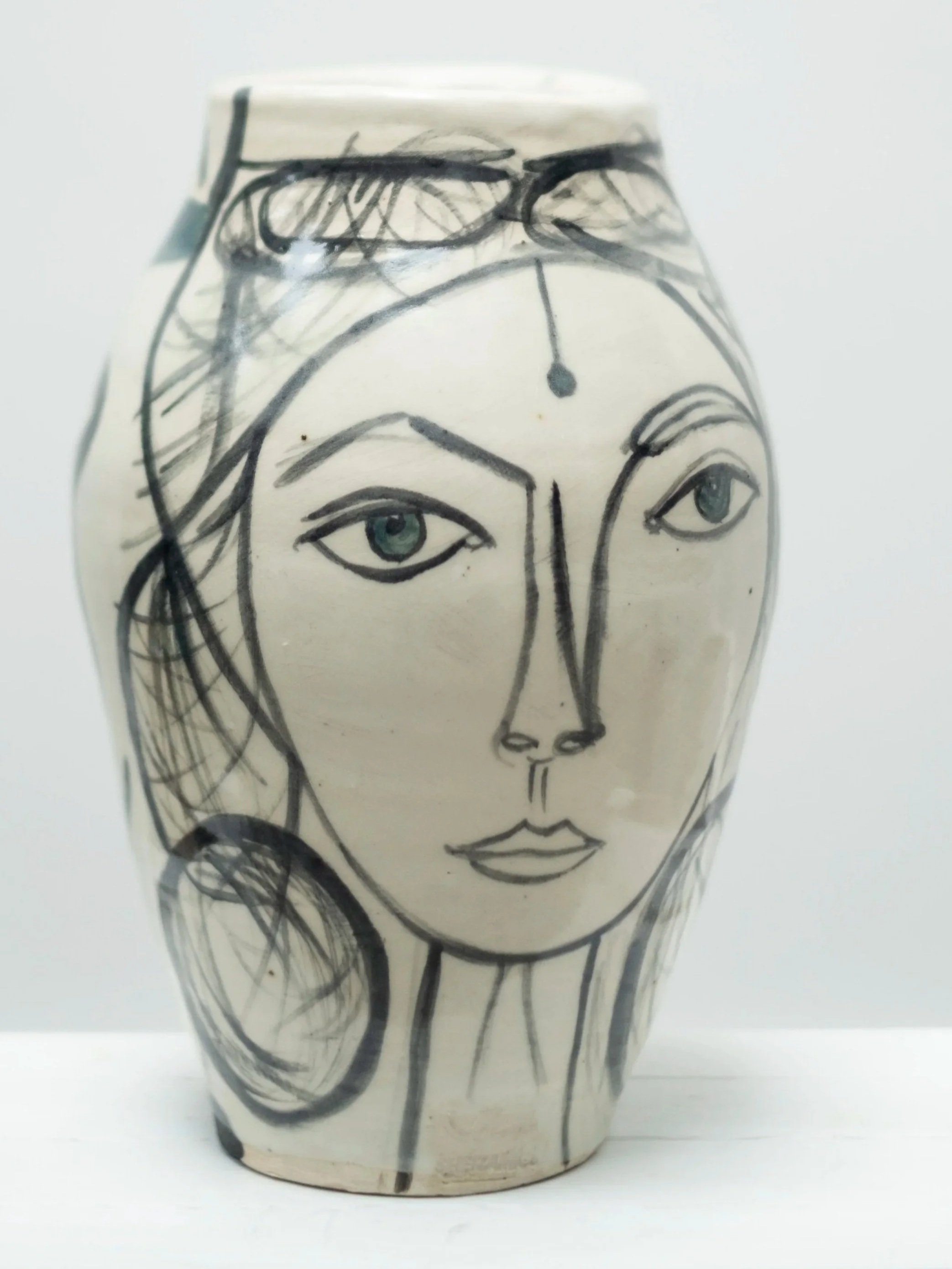 Ceramic vase with a black abstract sketch of a woman's face, including eyes, nose, lips, and hair, with glasses resting on top of a headband.