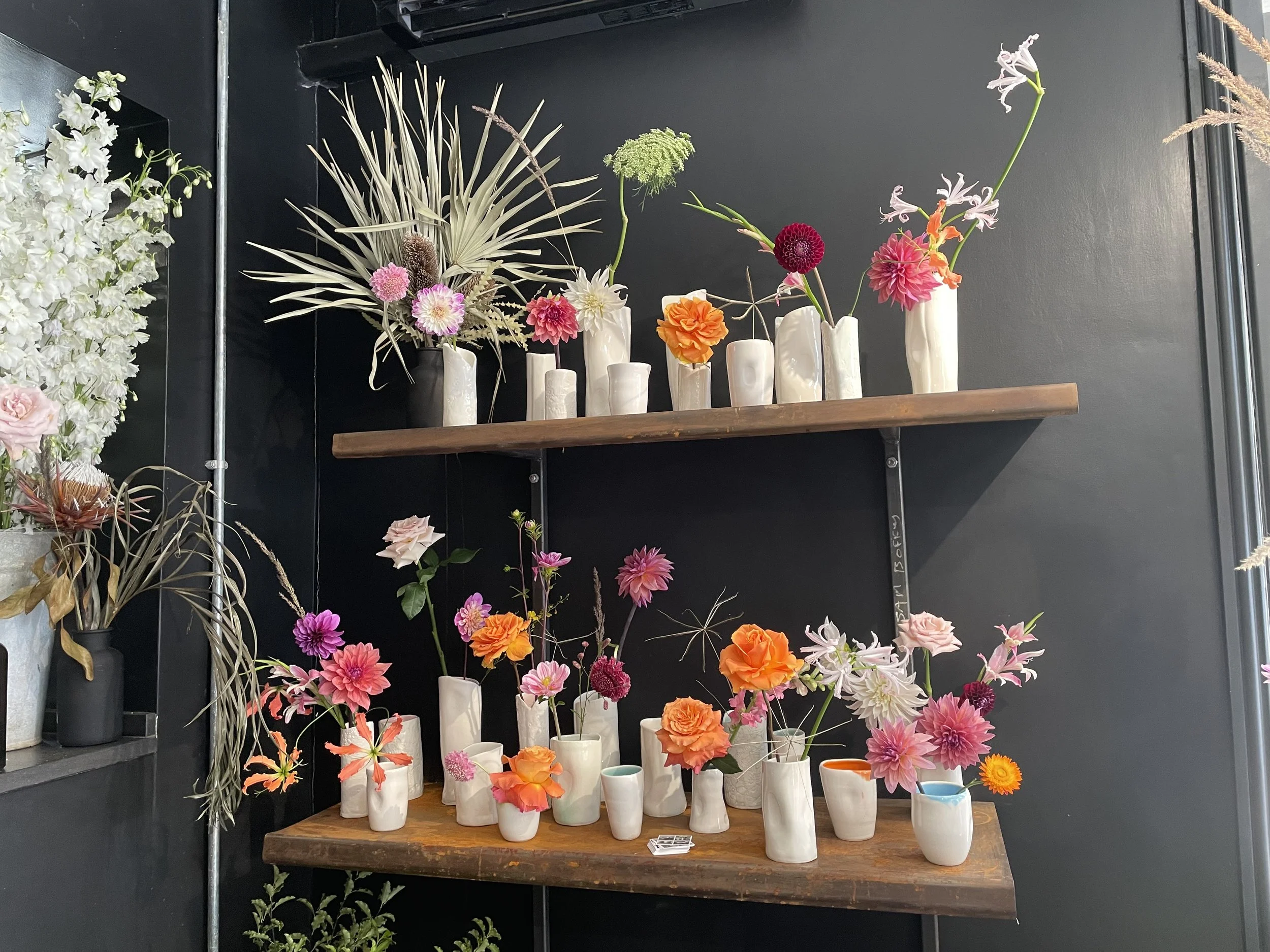 Display of colorful flowers in white vases on wooden shelves against a black wall.