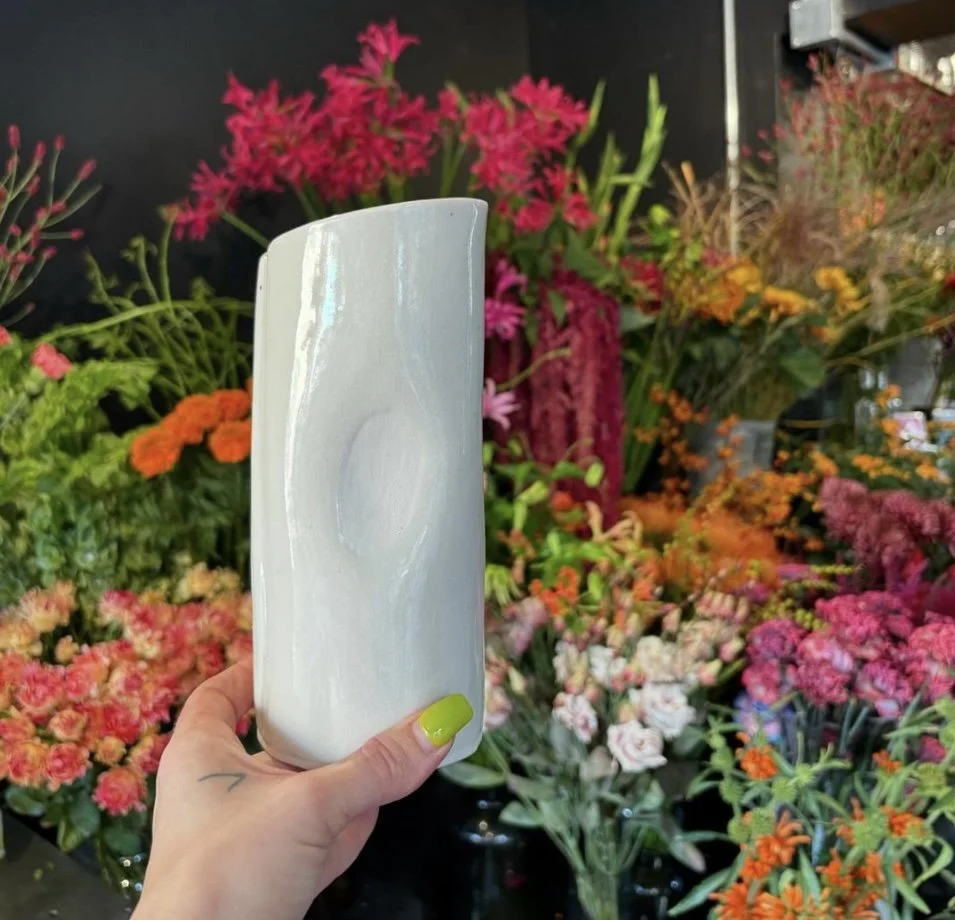 A hand holding a white ceramic vase in front of a colorful display of various flowers including pink, orange, and purple blossoms.