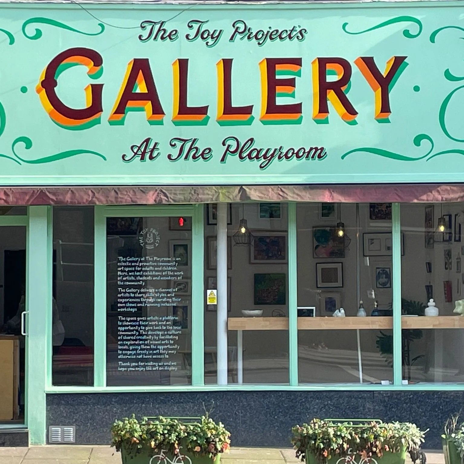 Bright green storefront sign for 'The Toy Projects Gallery at the Playroom' with large, colorful text and decorative swirls, with a large window display showing framed artwork inside and potted flowers outside.