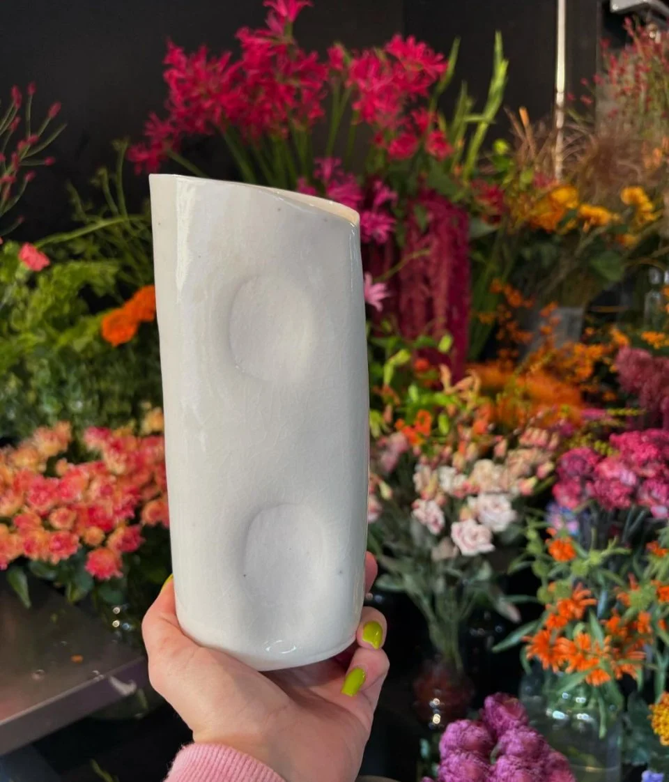 A white candle held by a person with neon green painted fingernails, in front of a colorful array of pink, orange, and purple flowers.