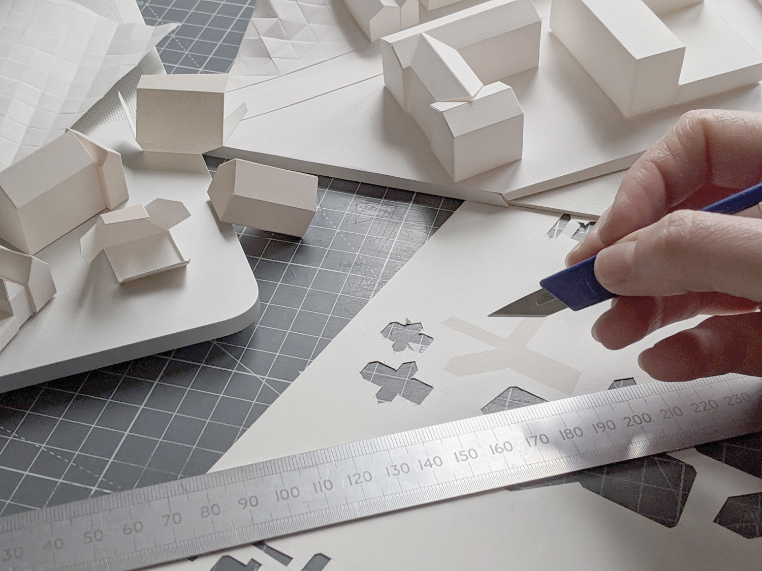 Small paper models being constructed by a person at a desk with scalpel and ruler in hand