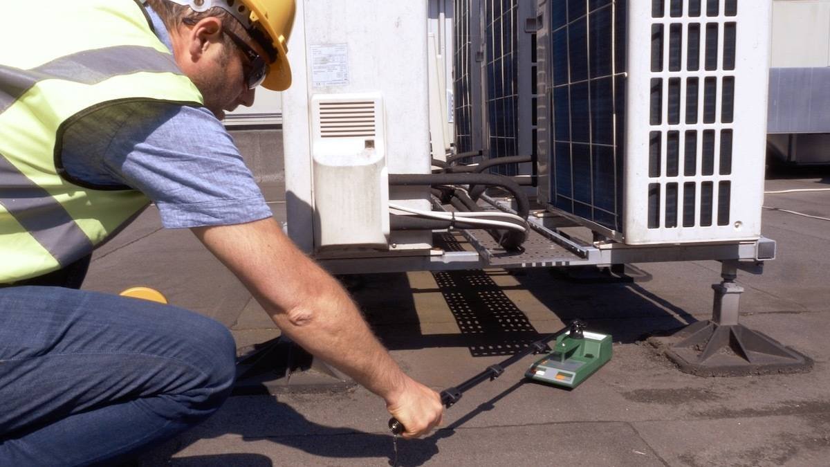 A worker wearing a yellow safety helmet and vest crouches on a rooftop, using a radiation detector to check the ground near HVAC units.