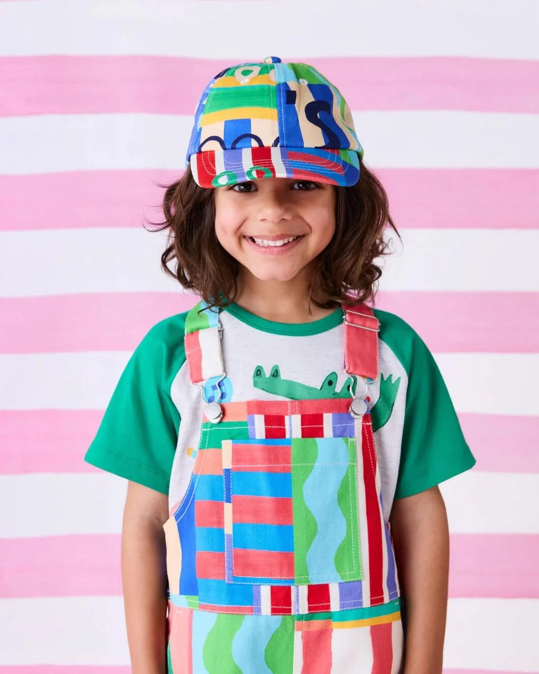 young girl standing in front of light pink striped background wearing colorful hat, shirt, and overalls