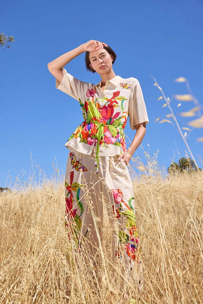 woman standing in brush wearing a floral silk shirt and pants shielding her eyes from the sun