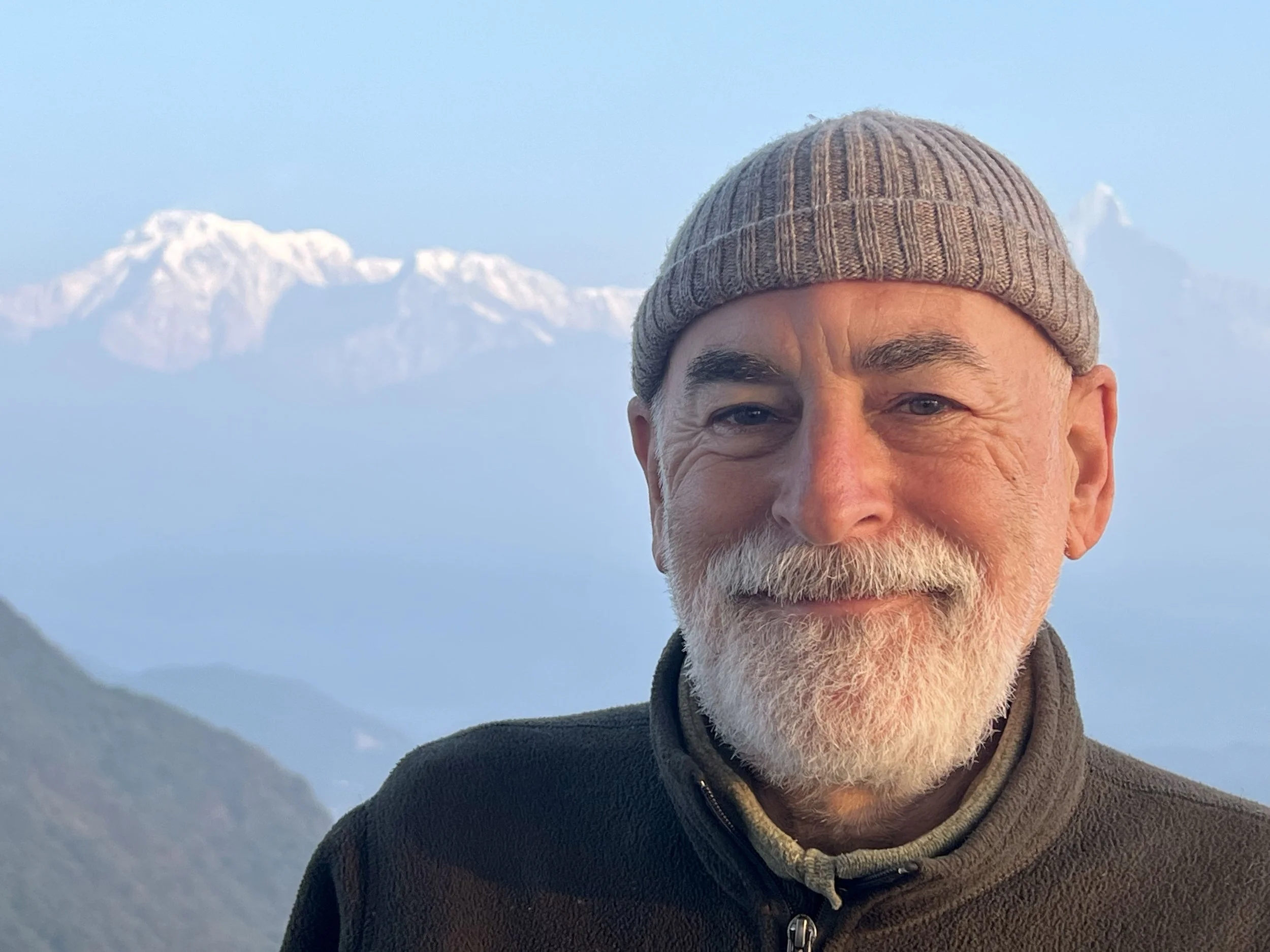 A smiling older man with a white beard and mustache, wearing a gray beanie and a dark jacket, outdoors with snow-capped mountains in the background.