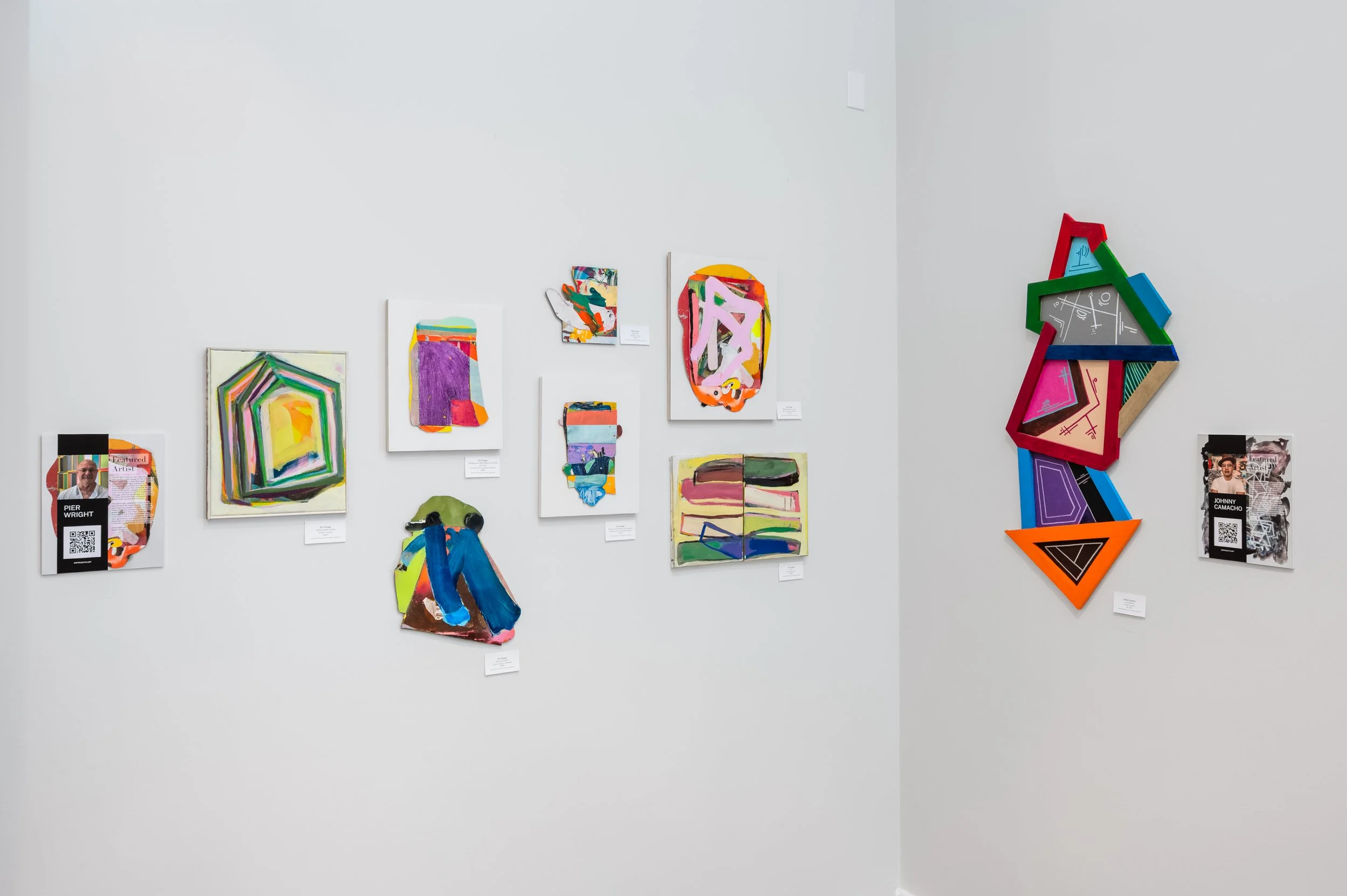 Colorful abstract art pieces and collages displayed on gallery walls, including paintings, paper collages, and three-dimensional geometric art. There are labels and QR codes next to some artworks.