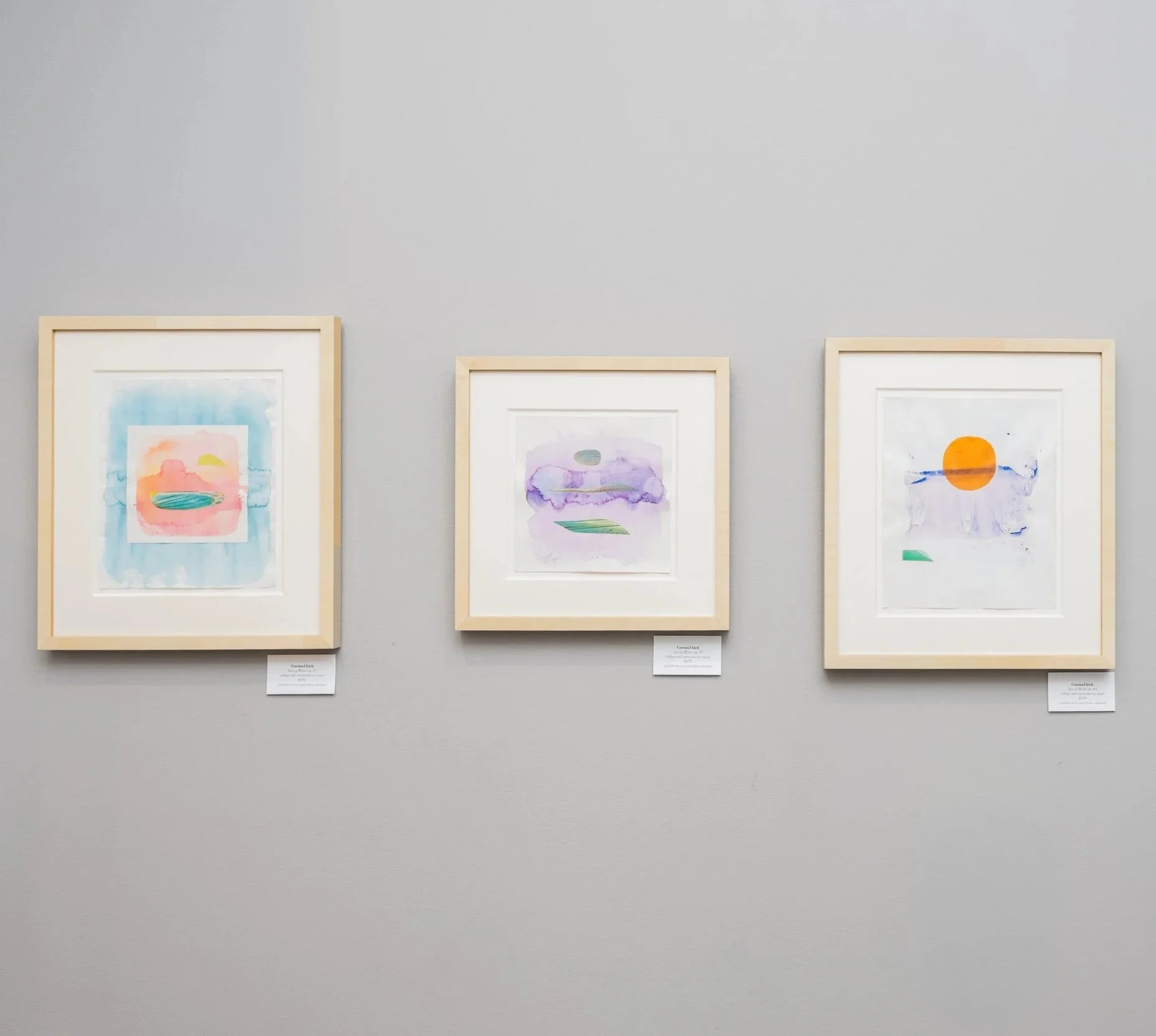 Three framed watercolor paintings on a gray gallery wall, each featuring abstract landscapes with pastel and bright colors.