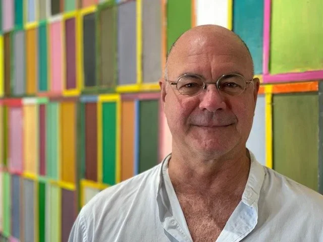 A man with glasses and a white shirt standing in front of a colorful grid wall.