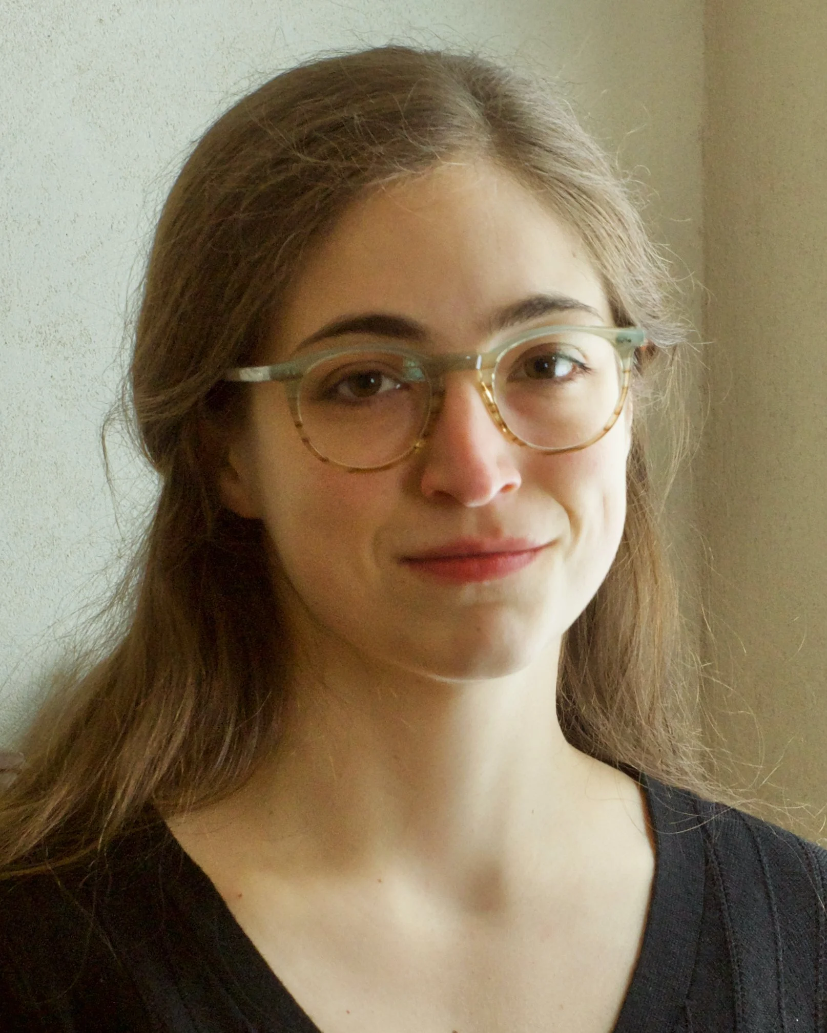 A young woman with long brown hair, wearing glasses and a black shirt, standing near a beige wall.