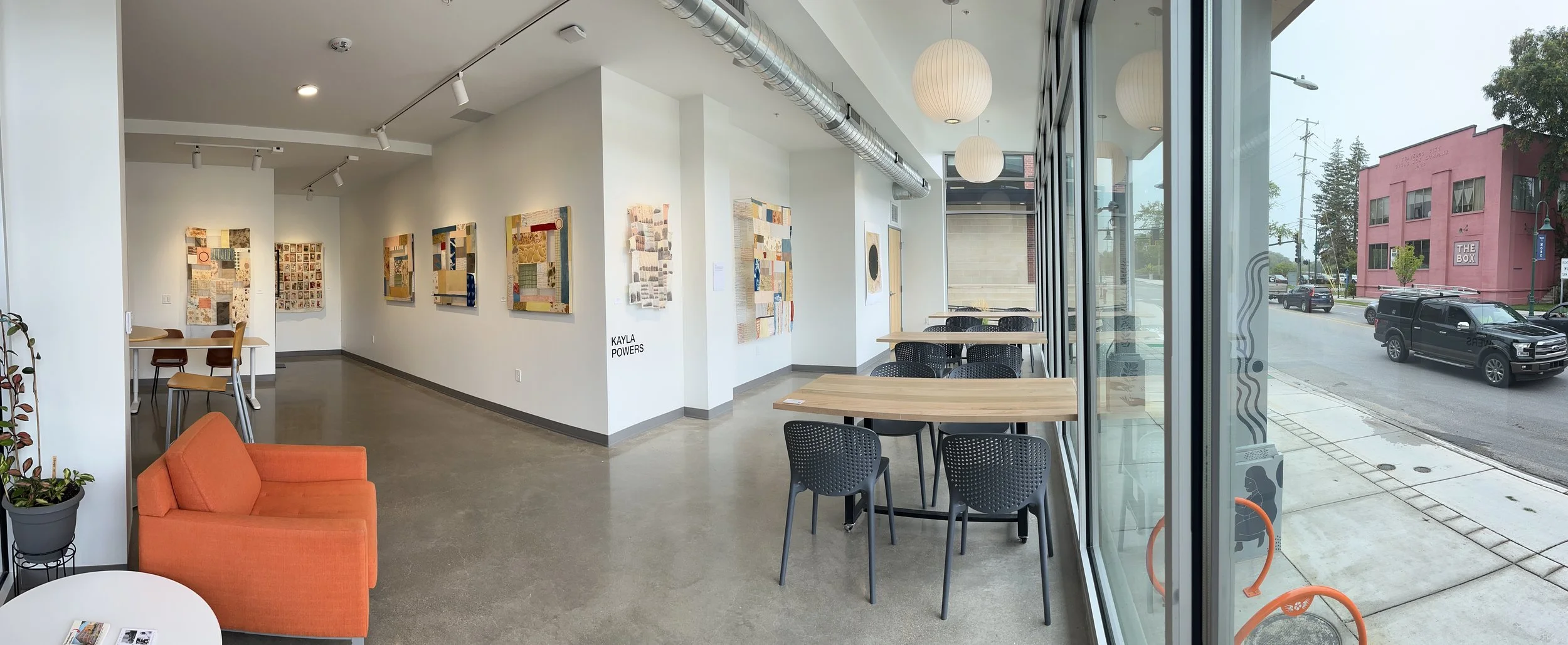 Interior of a modern art gallery with wall-mounted fabric art pieces, small sitting area with an orange sofa, and large windows overlooking parked cars and a pink building outside.