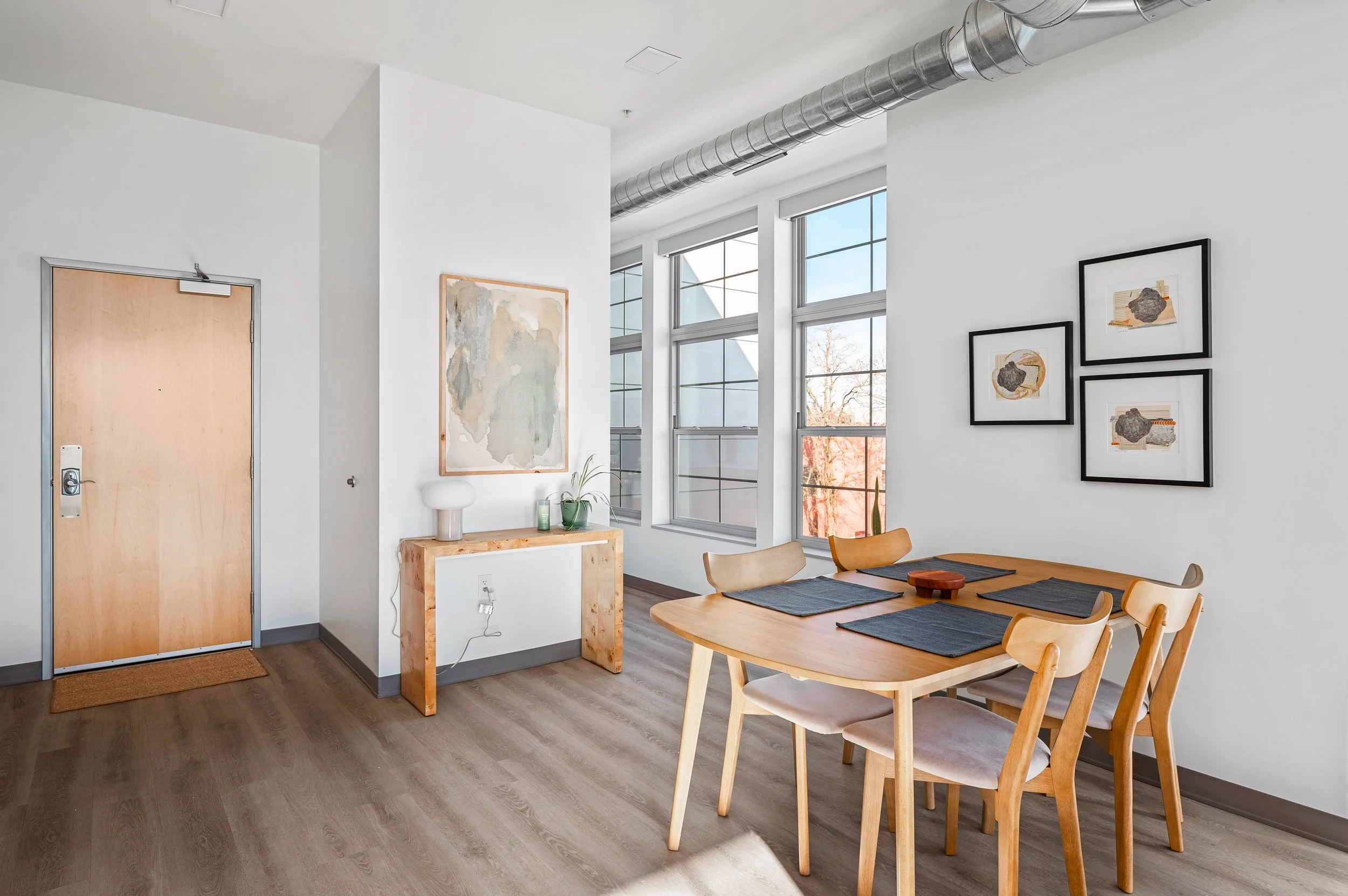 Modern dining area with wooden table set with four place mats, surrounded by light wooden chairs, against white walls decorated with framed artwork, large windows letting in natural light, and a wooden console with decorative items, in a contemporary apartment.