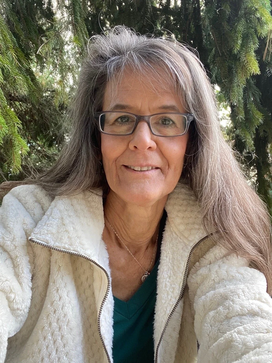 A middle-aged woman with long gray hair and glasses smiling outdoors in front of green trees, wearing a cream-colored fleece jacket and a dark shirt.