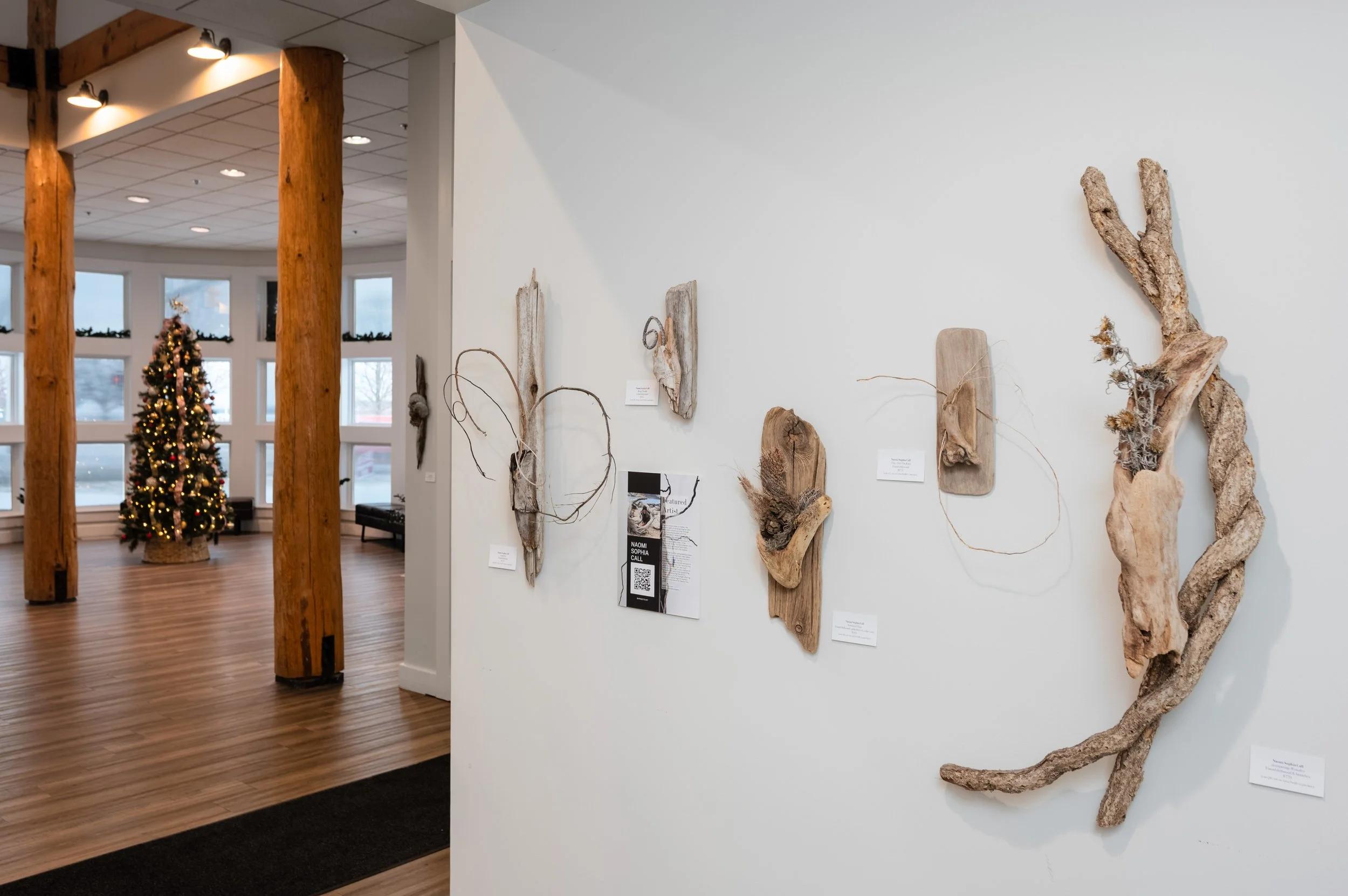 Art installation featuring driftwood pieces arranged on a white wall in a gallery, with a decorated Christmas tree visible in a room with wooden columns in the background.