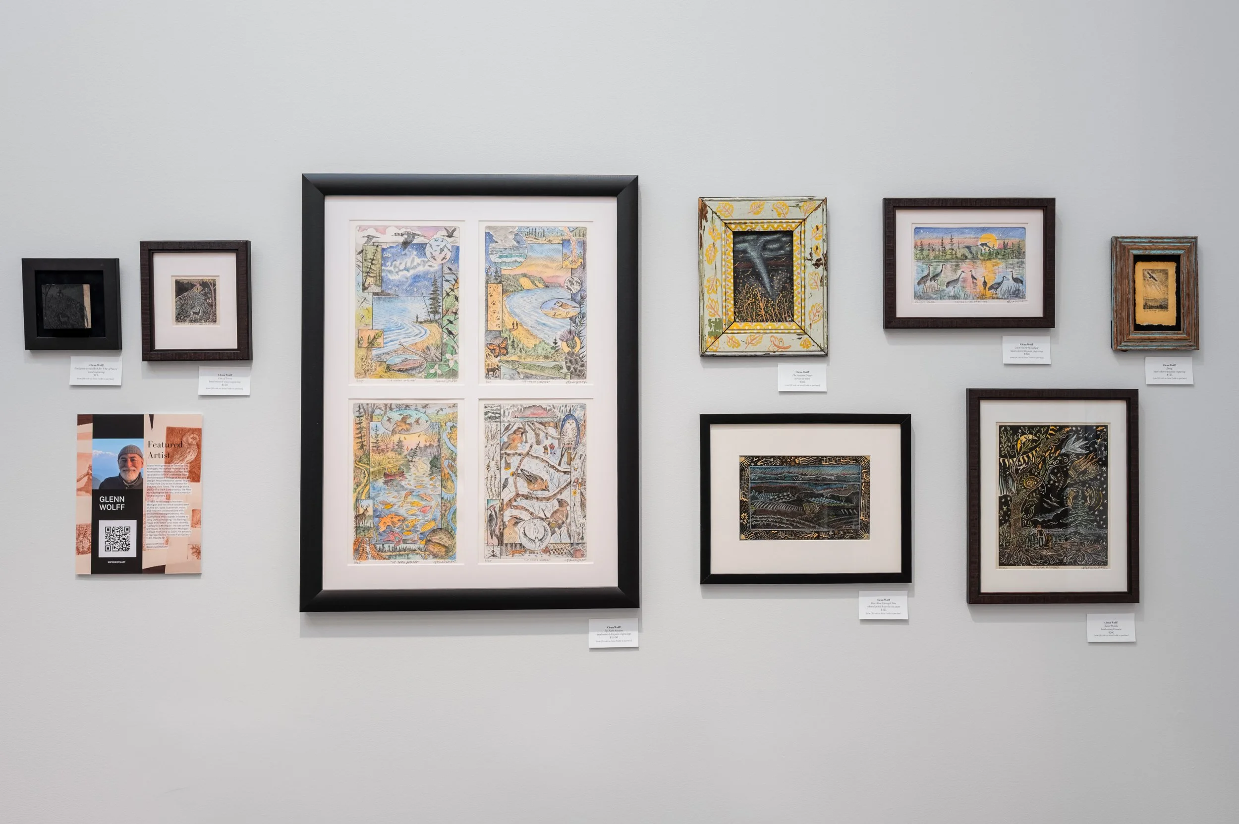 Wall display of various framed artworks, including colorful paintings, drawings, and prints, with a small information card and an artist profile featuring a QR code.