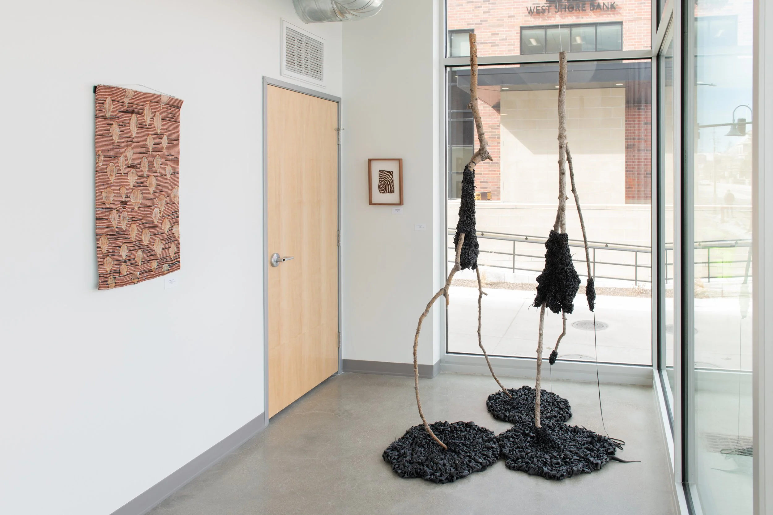 Art gallery interior displaying a textile wall piece on the left and two mixed media sculptures resembling tall slender figures made from branches and black textured material, positioned in front of large glass windows.