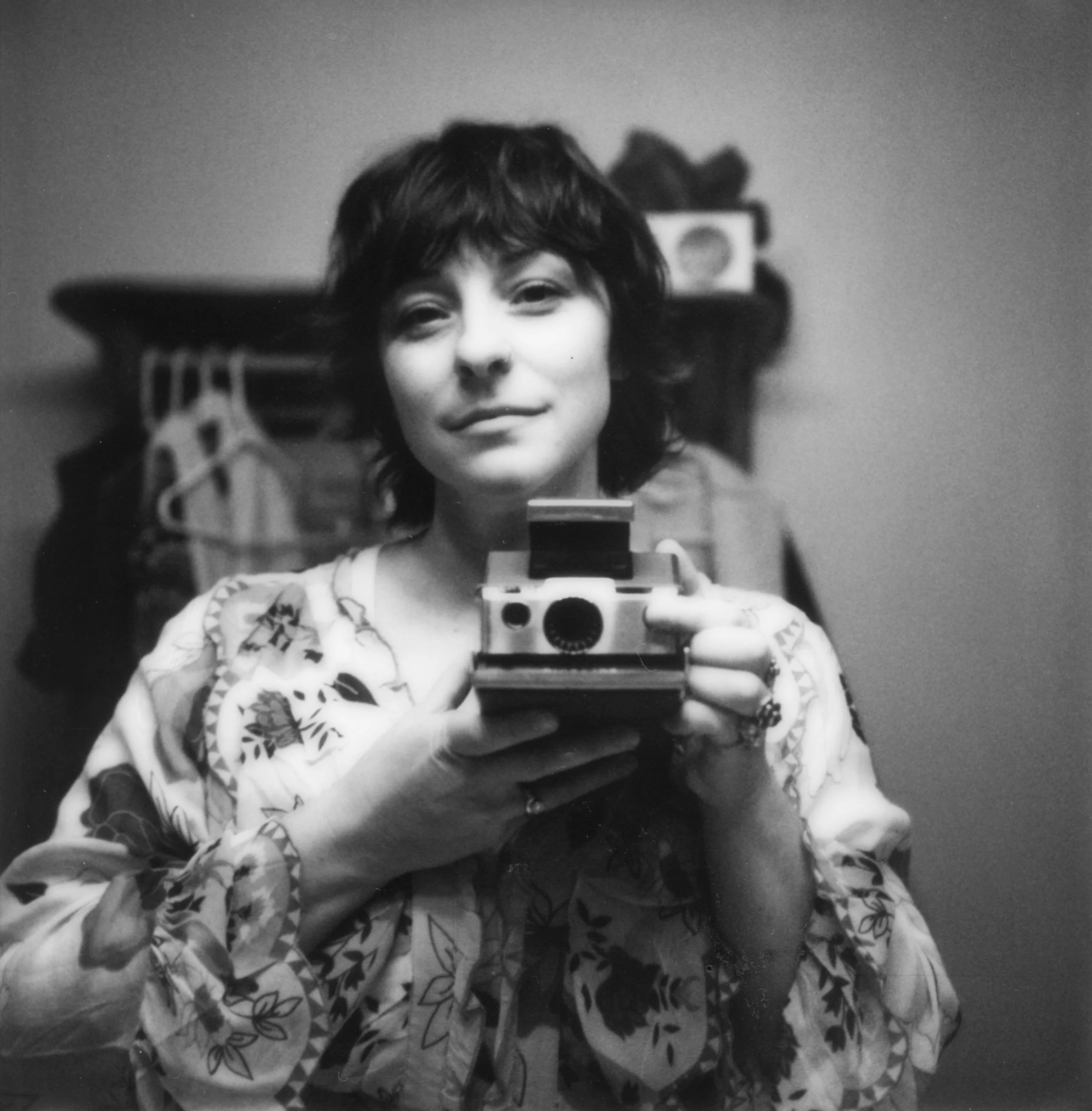 A woman with short dark hair taking a selfie in front of a mirror, holding a camera, with a blurred background showing a dresser and personal items.