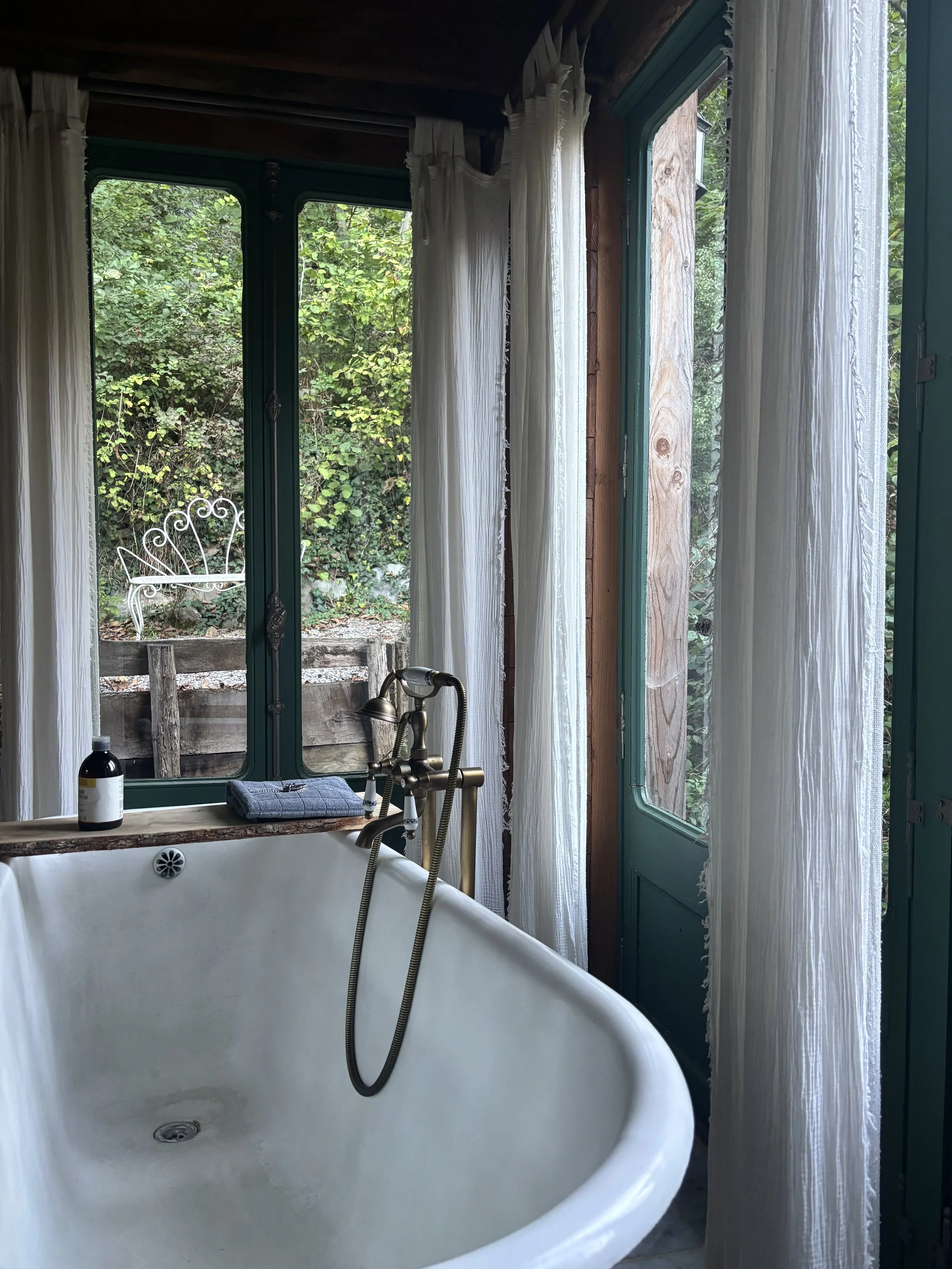 A rustic bathroom with a free-standing bathtub, brass fixtures, and large windows with white curtains, overlooking greenery outside.