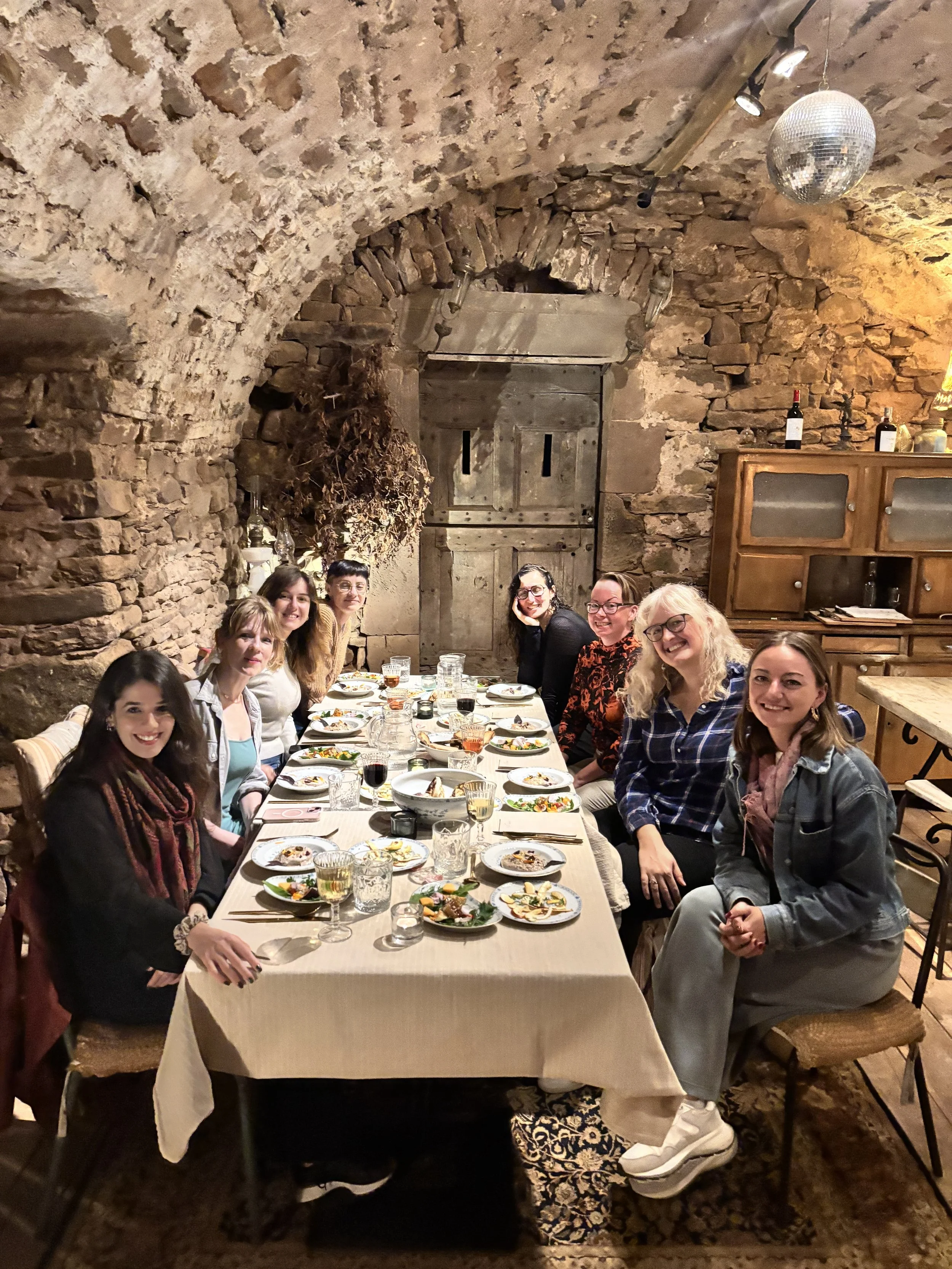 A group women sitting around a dining table in a rustic brick-walled cellar, enjoying a meal with plates of food, glasses of wine, and drinks, under soft lighting with a disco ball hanging from the ceiling.