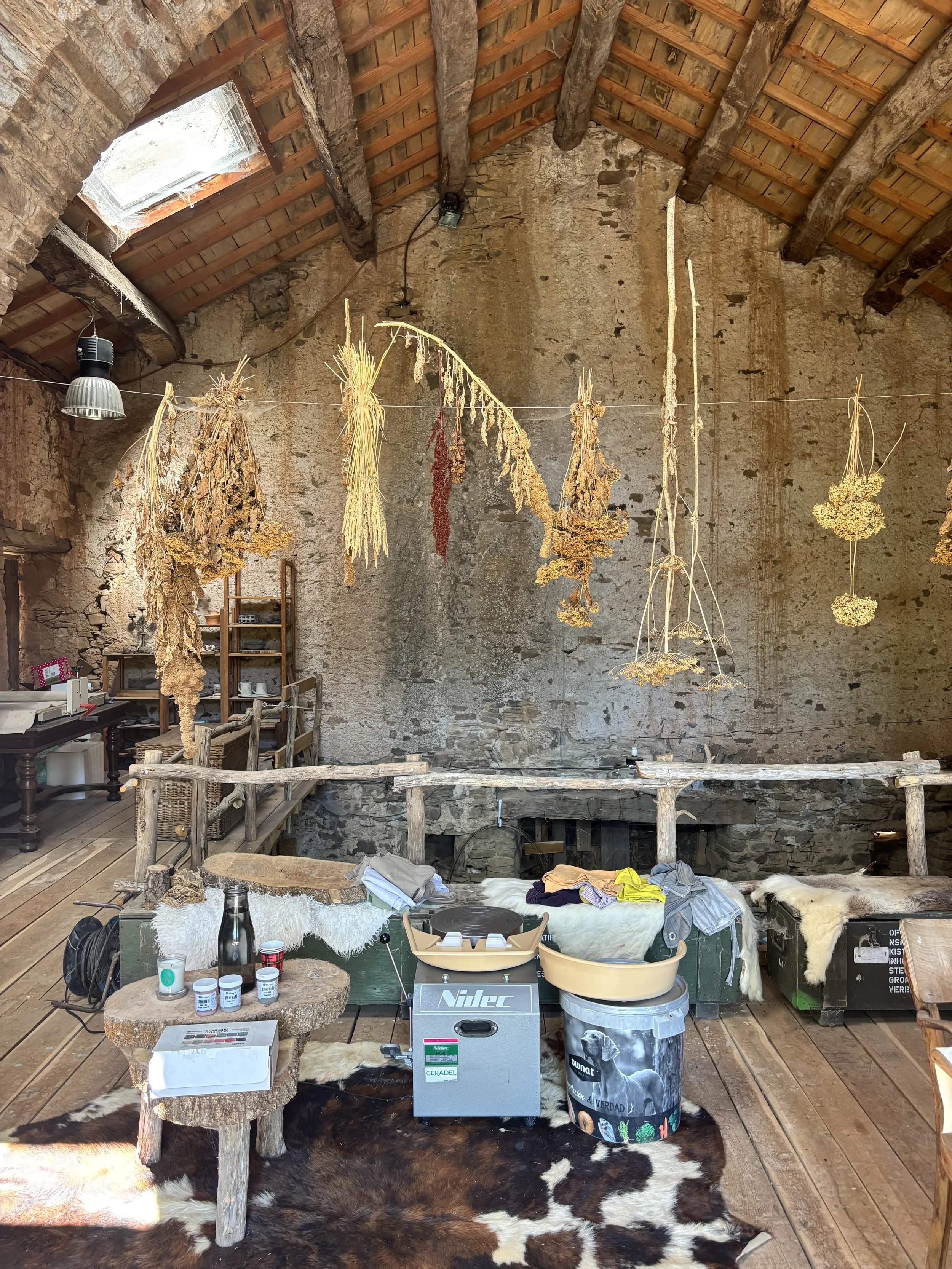 Interior of a rustic room with hanging dried herbs and flowers, a stone wall, wooden ceiling with exposed beams, and a cowhide rug on the wooden floor.