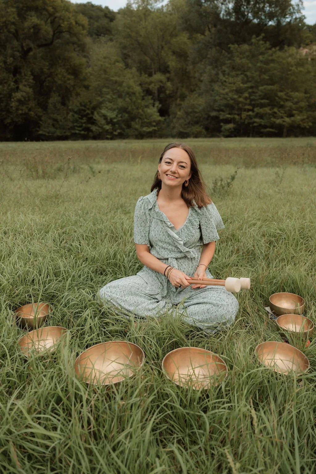 A woman sitting cross-legged in a grassy field surrounded by singing bowls, holding a mallet, smiling, with a forested background.