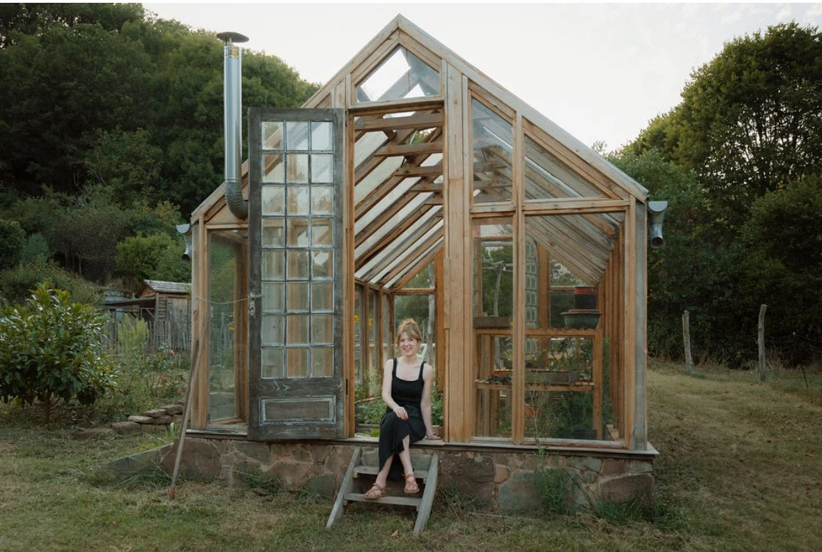 A young woman in a black dress sitting in front of a small wooden greenhouse with glass panels, surrounded by greenery.