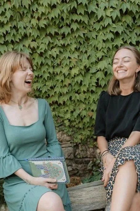 Two women laughing and enjoying each other's company outdoors, sitting on a wooden bench in front of a lush green ivy-covered wall.