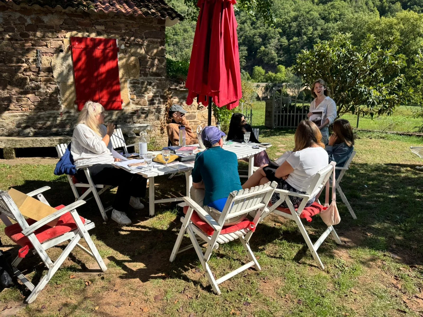 A group of women gathered outdoors around a white table with books, notebooks, and glasses of water, listening to a woman standing and speaking in front of a stone building with a red window shutter, red umbrella, green trees, and grassy area.