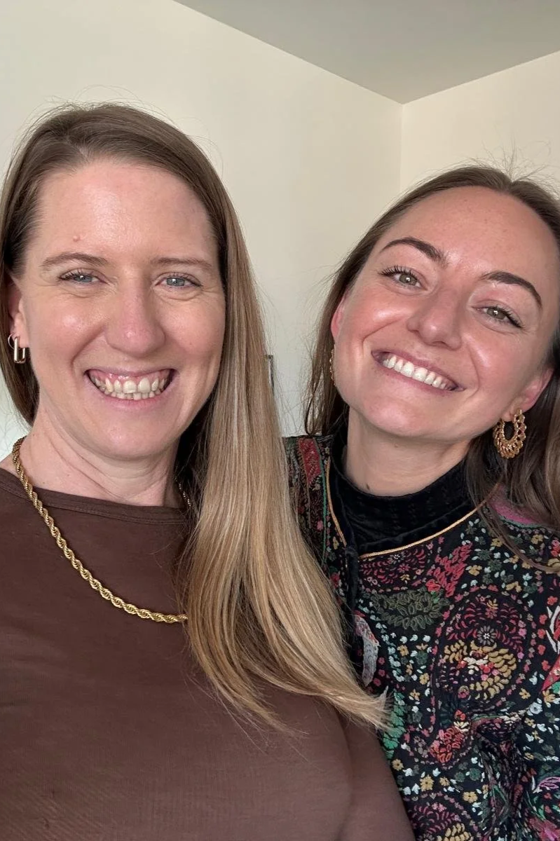 Two women smiling for a close-up photo indoors with a plain background.