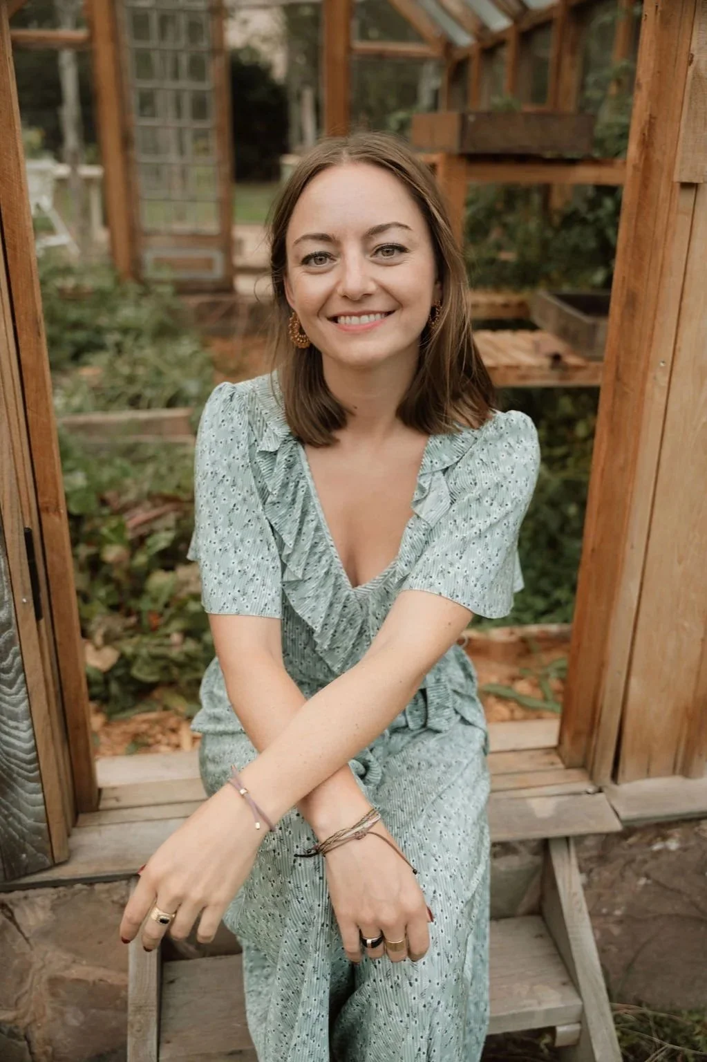 A woman with shoulder-length brown hair, smiling, sitting inside a wooden structure in a garden or greenhouse, wearing a light blue, patterned dress, jewelry, and rings.