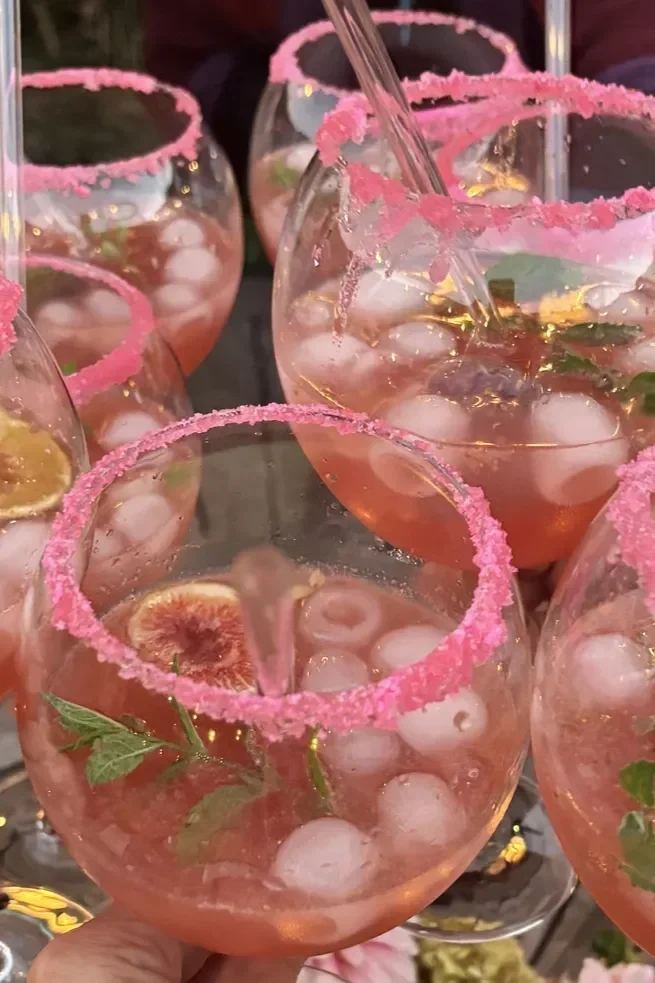 Pink-colored cocktails in large wine glasses with pink sugar rims, ice, slices of fig, and mint leaves, garnished for a festive occasion.