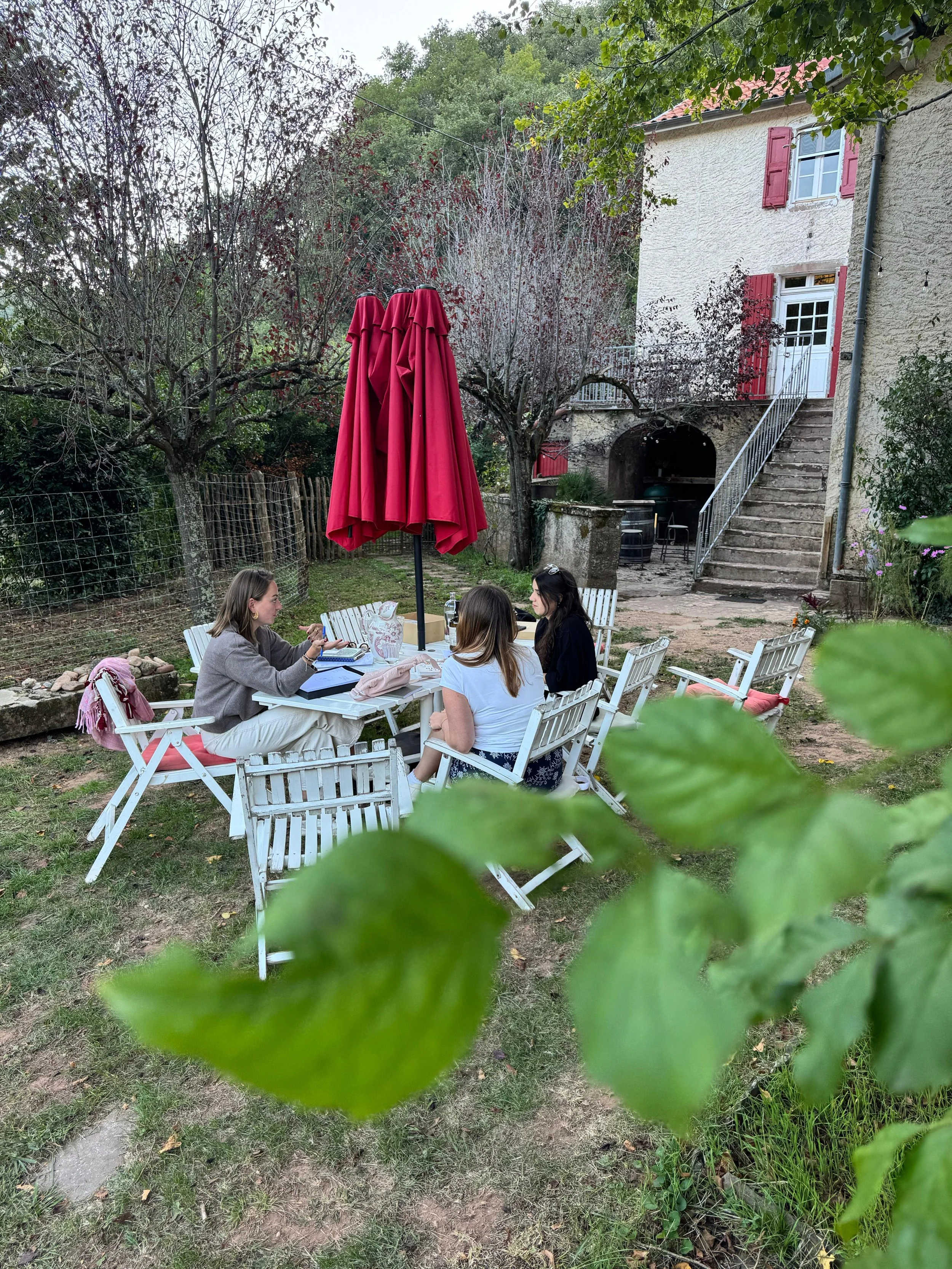 Three women sit around a white outdoor table with a large red umbrella in a garden, adjacent to a stone house with red shutters and a staircase. The background includes trees and a wire fence.