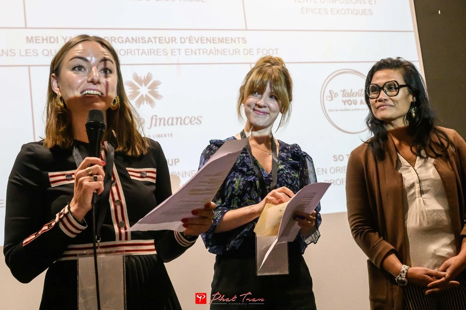 Three women standing on stage during a presentation or conference, with a large screen displaying text behind them. The woman on the left is speaking into a microphone, holding papers, wearing a black dress with red and white striped accents. The woman in the middle has reddish-brown hair, holding papers, wearing a dark floral blouse. The woman on the right has dark hair, glasses, wearing a brown jacket over a beige top.