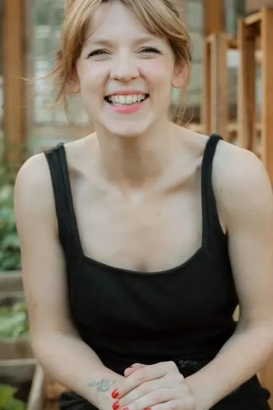 A woman with red hair and a black tank top smiling outdoors, with a greenhouse or garden in the background.