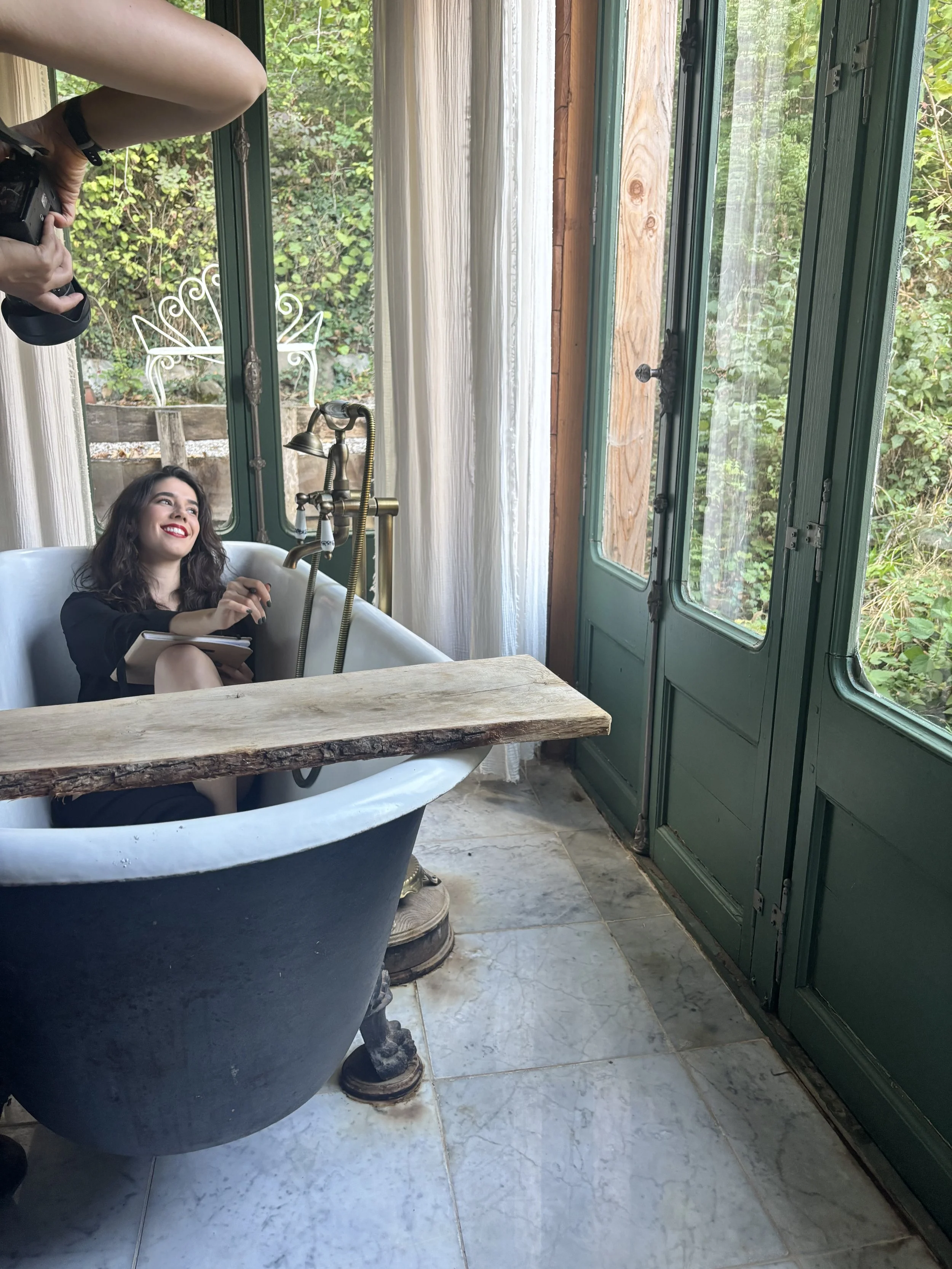 A woman with dark hair and red lipstick sitting in a vintage clawfoot bathtub with a wooden plank across the top, holding a notebook and pen, while a photographer photographs her inside a room with large green windows and a garden outside.