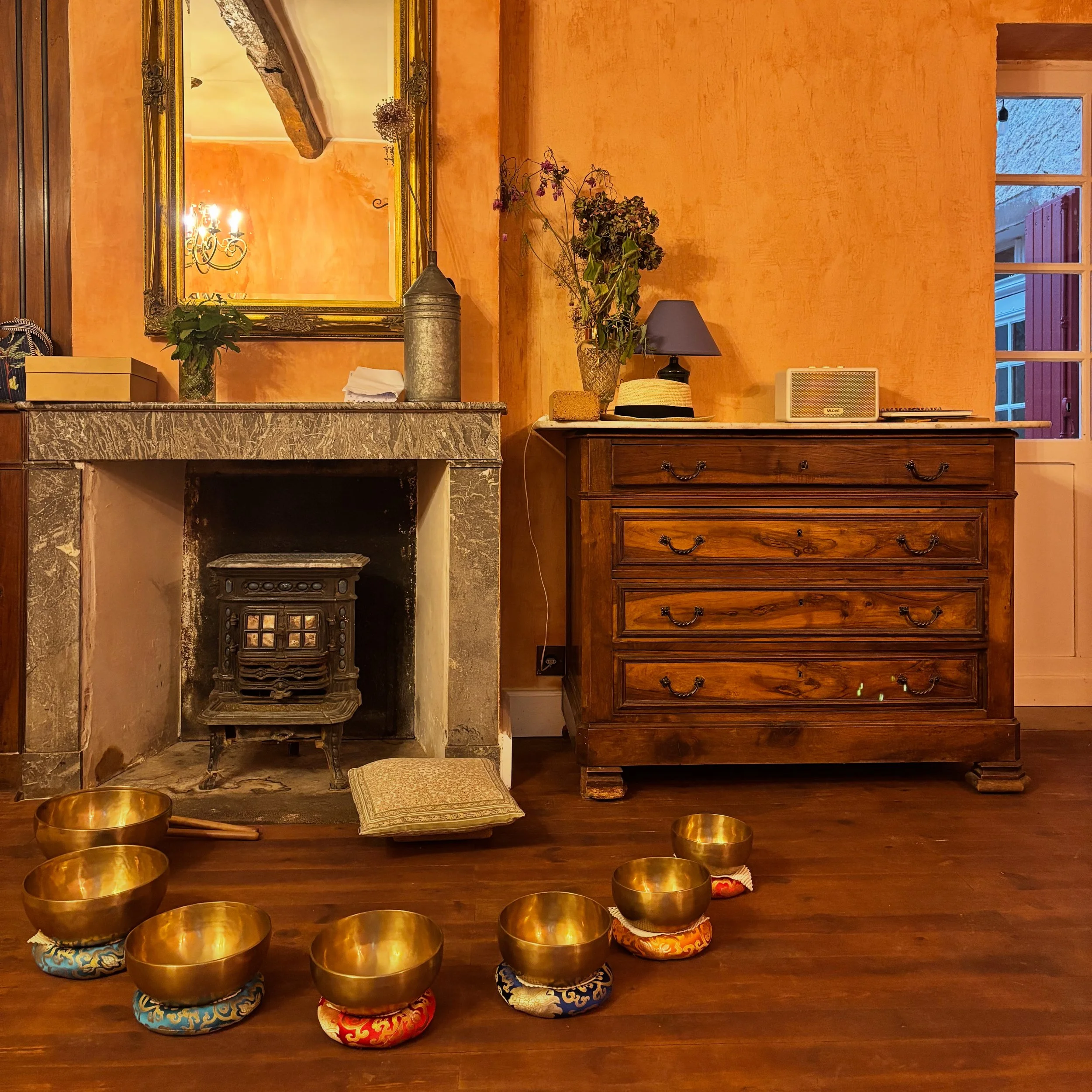Living room corner with a fireplace, a large mirror above it, a wooden chest of drawers with decorative items on top, and a window showing an outdoor view. Golden singing bowls with pillows in front of the fireplace.