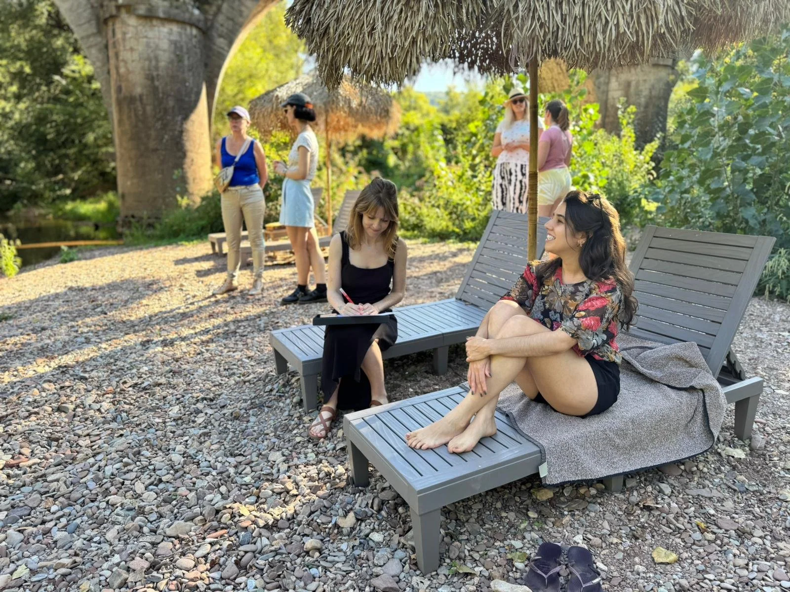 A woman sitting on a lounge chair with her legs crossed and smiling, another woman sitting nearby writing in a notebook, under a thatched umbrella on a pebbled beach with others standing and talking in the background.