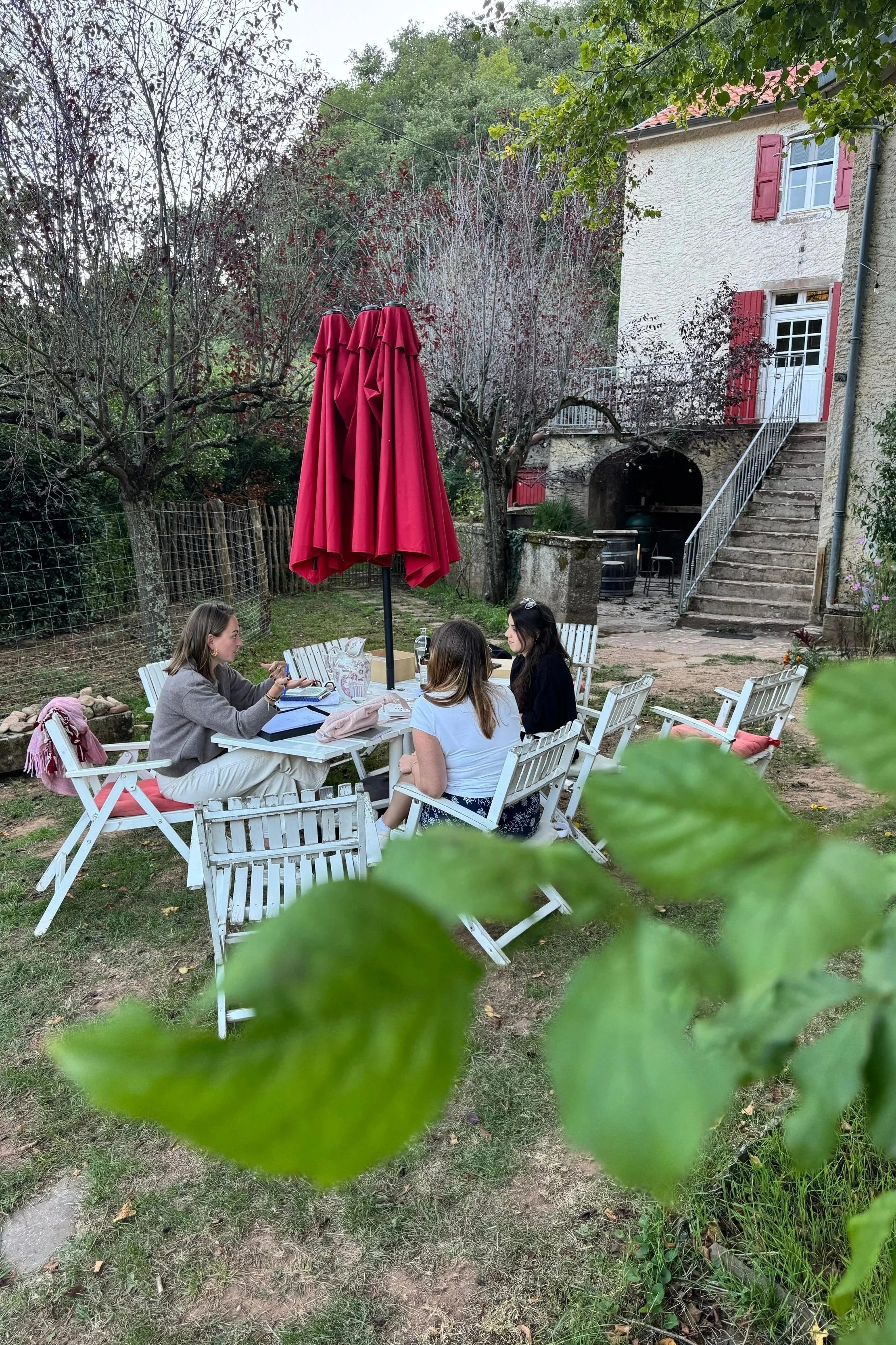 Three women sitting at a white outdoor table with a red umbrella in a garden, engaging in conversation. The garden has trees, a stone house with red shutters, and a staircase leading to an upper level.