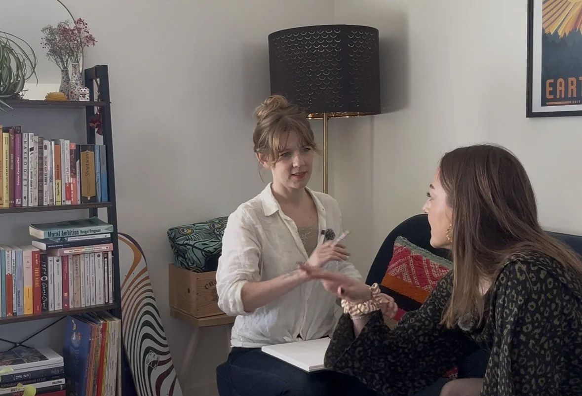 Two women are sitting in a living room, one handing a band to the other. There is a bookshelf filled with books and a skateboard leaning against the wall nearby. A lamp and framed poster are also visible in the background.