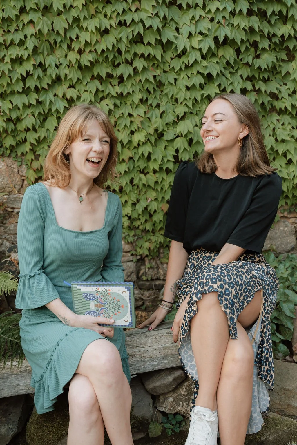 Two women sitting on a wooden bench outdoors, laughing and smiling at each other. They are surrounded by green ivy on a stone wall.
