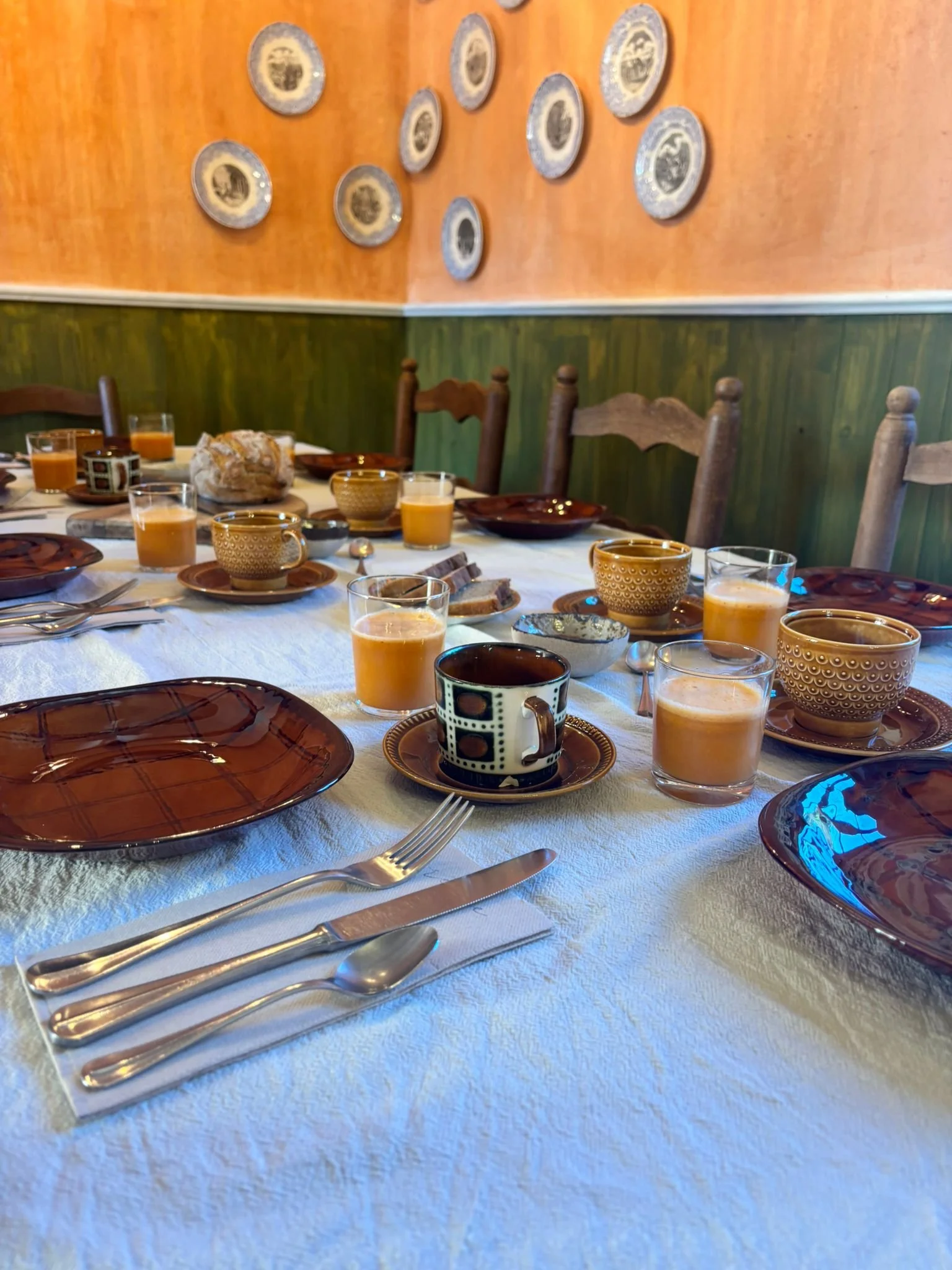 A dining table set with ceramic bowls, glass cups of orange juice, a black-and-white patterned mug, and a loaf of bread, with wooden chairs and a wall decorated with ceramic plates in the background.