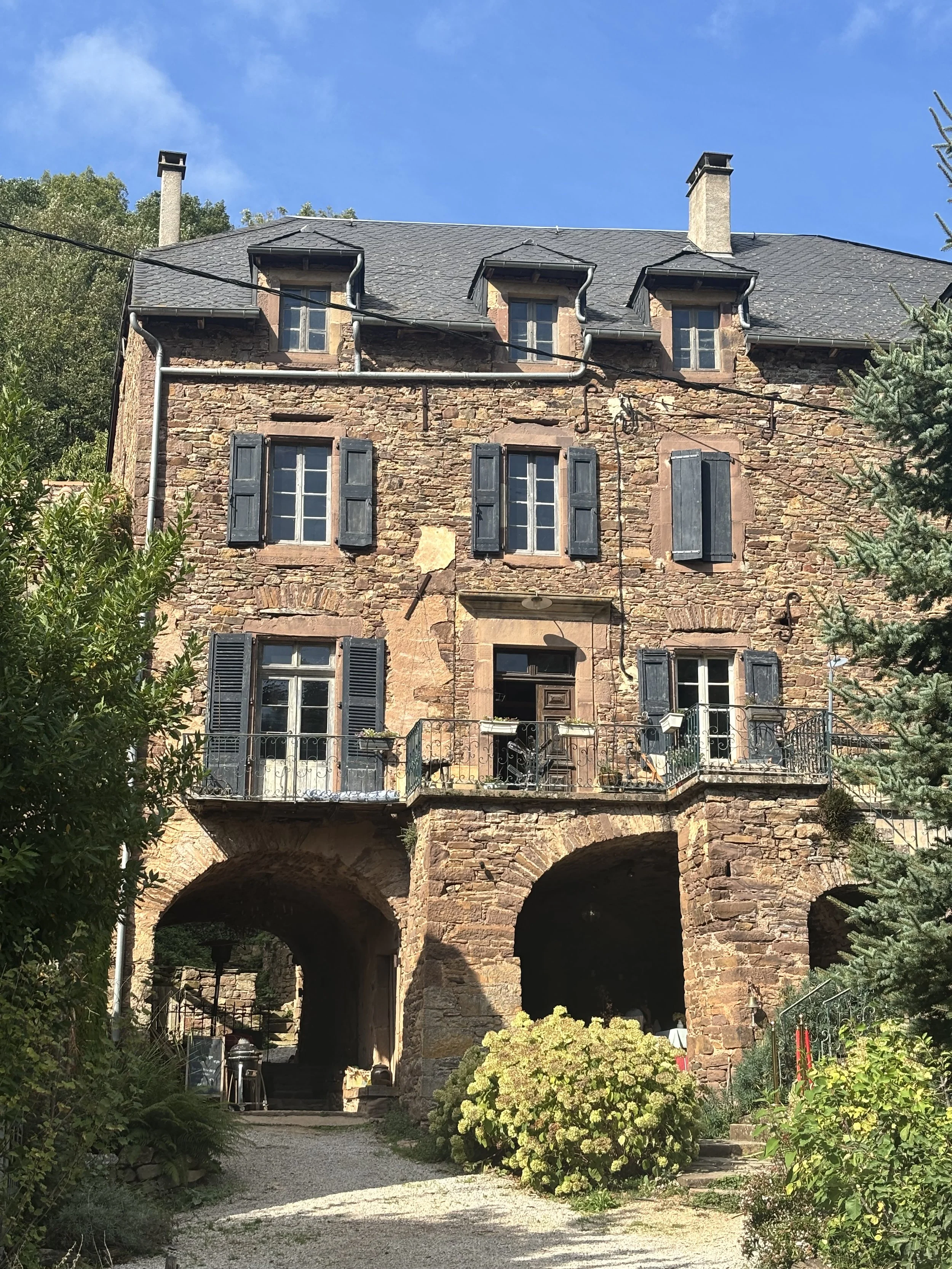 A stone house with multiple black window shutters, a balcony, and an arched stone base. There are plants and trees around the house, with a clear blue sky overhead.